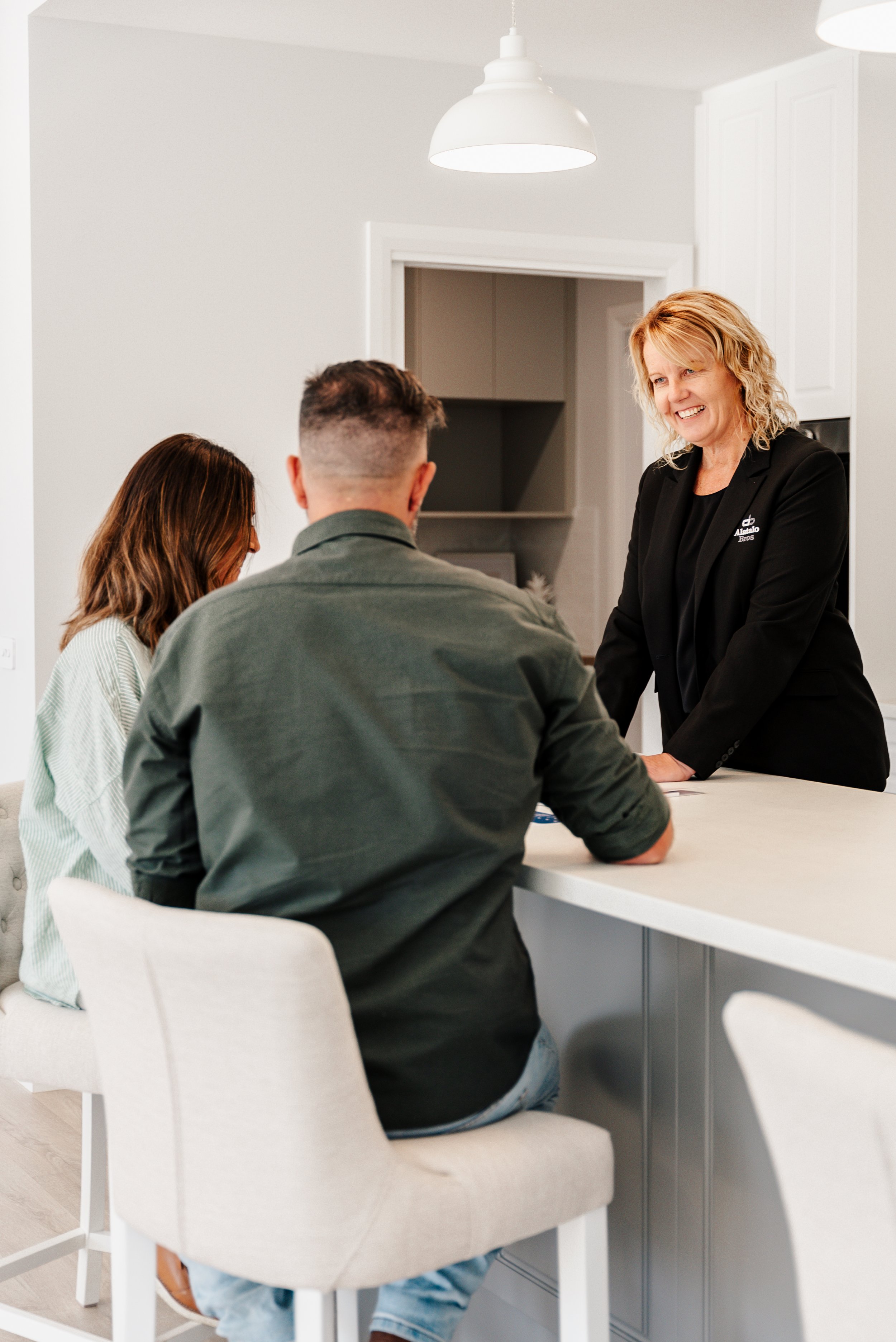 A real estate agent is smiling and talking with two prospective clients seated at a kitchen counter in a modern home. The room is well-lit with pendant lighting.