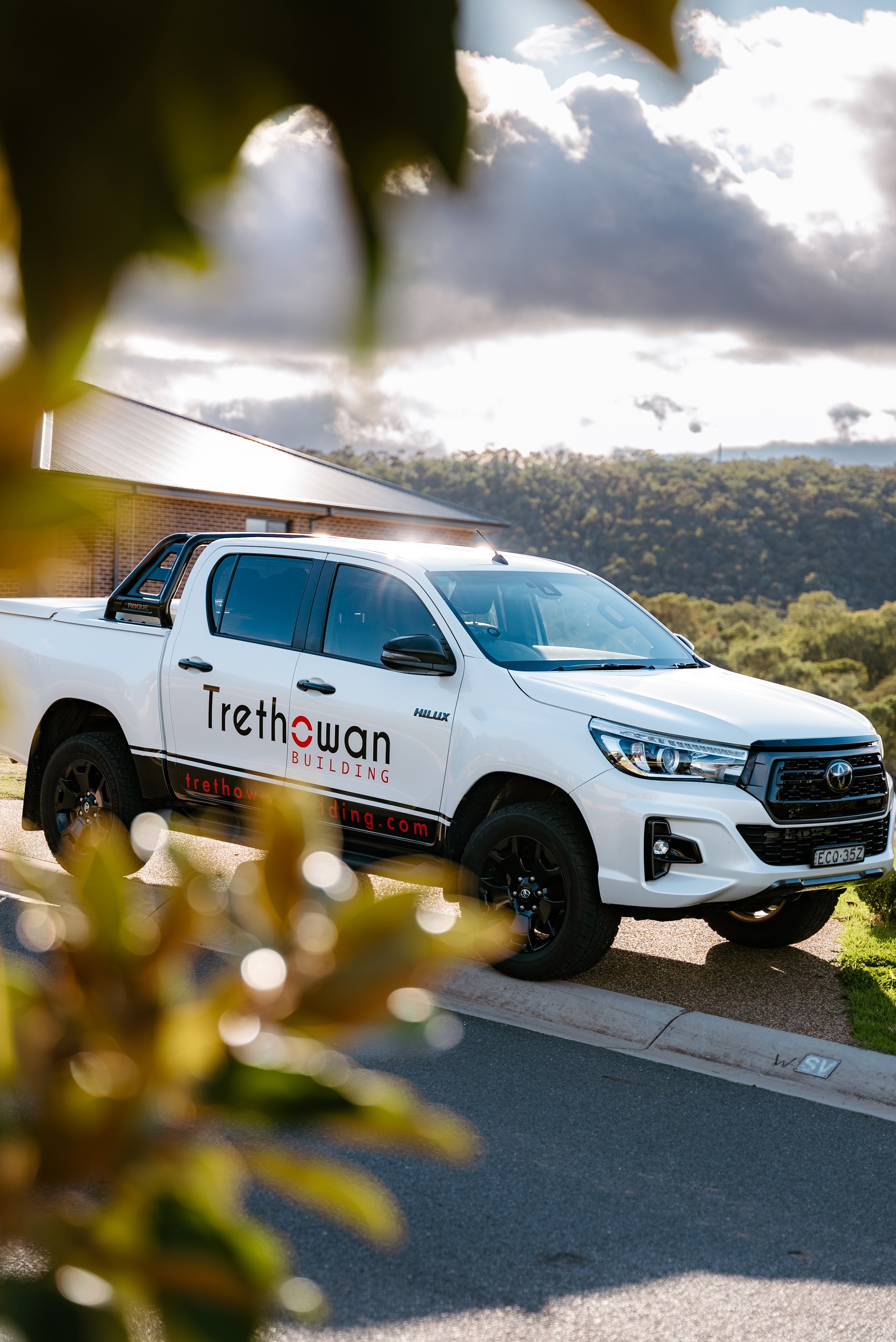 A white Toyota Hilux pickup truck with "Trethowan Building" logo parked beside a house, with trees and hills in the background.