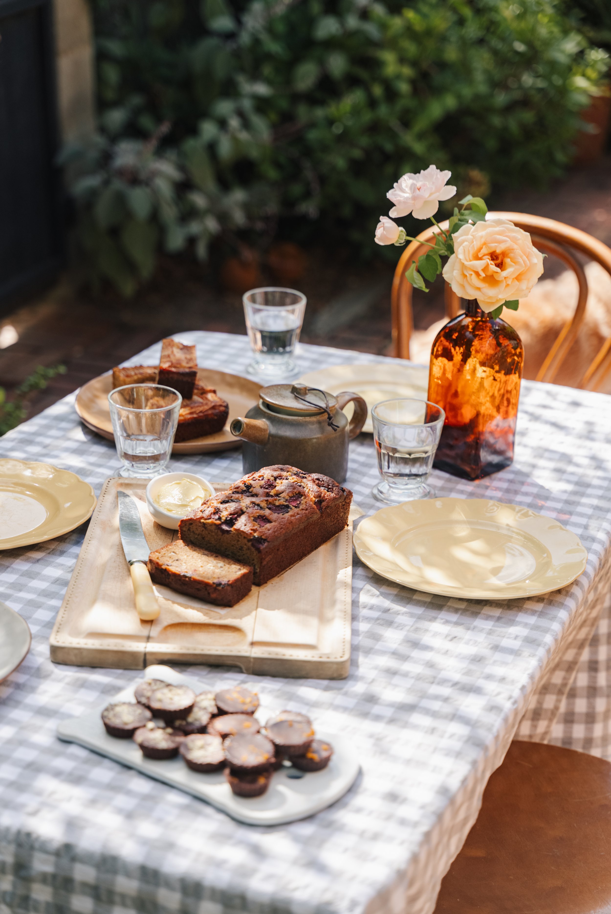 A table set for an outdoor meal with a loaf of bread, knife, plate of sliced bread, a teapot, glasses of water, small muffins, and a vase with roses on a checkered tablecloth.