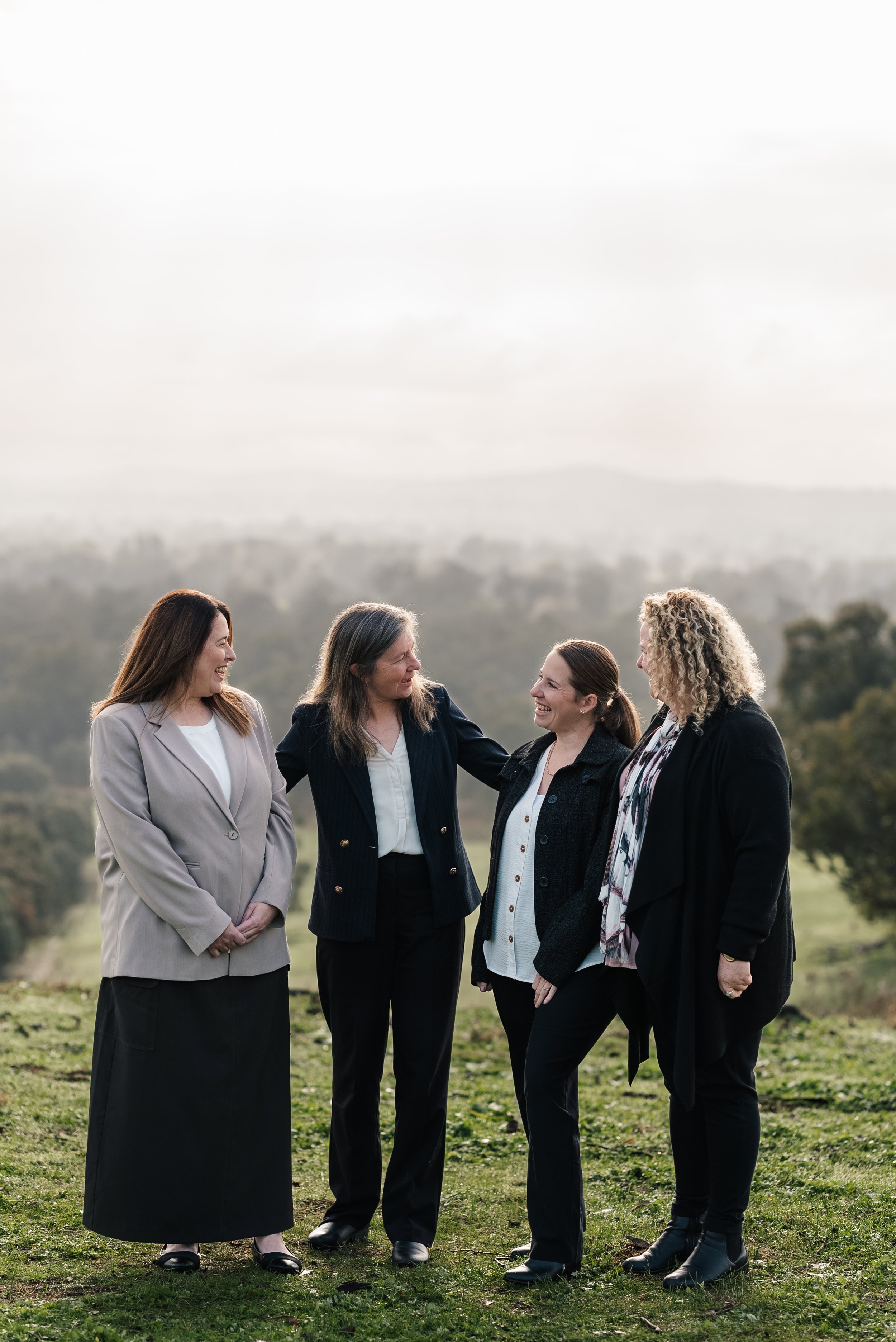 Four women standing outdoors, smiling and talking, with a scenic nature background.