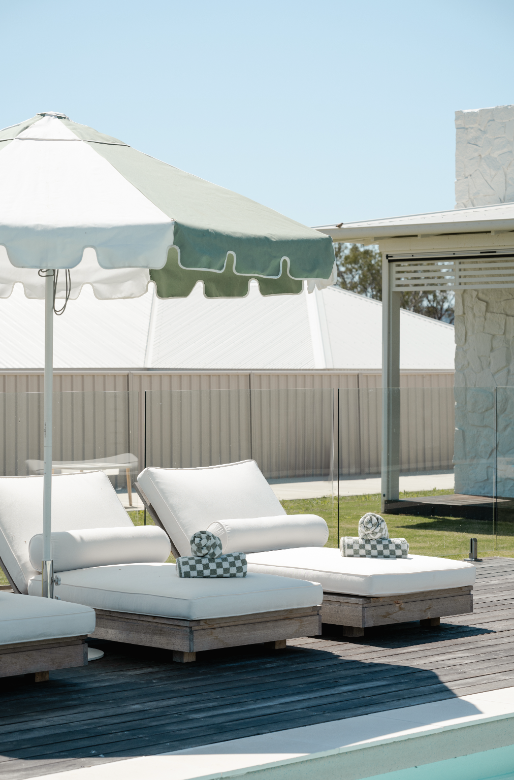 Outdoor poolside with white lounge chairs, green and white umbrella, and rolled towels.