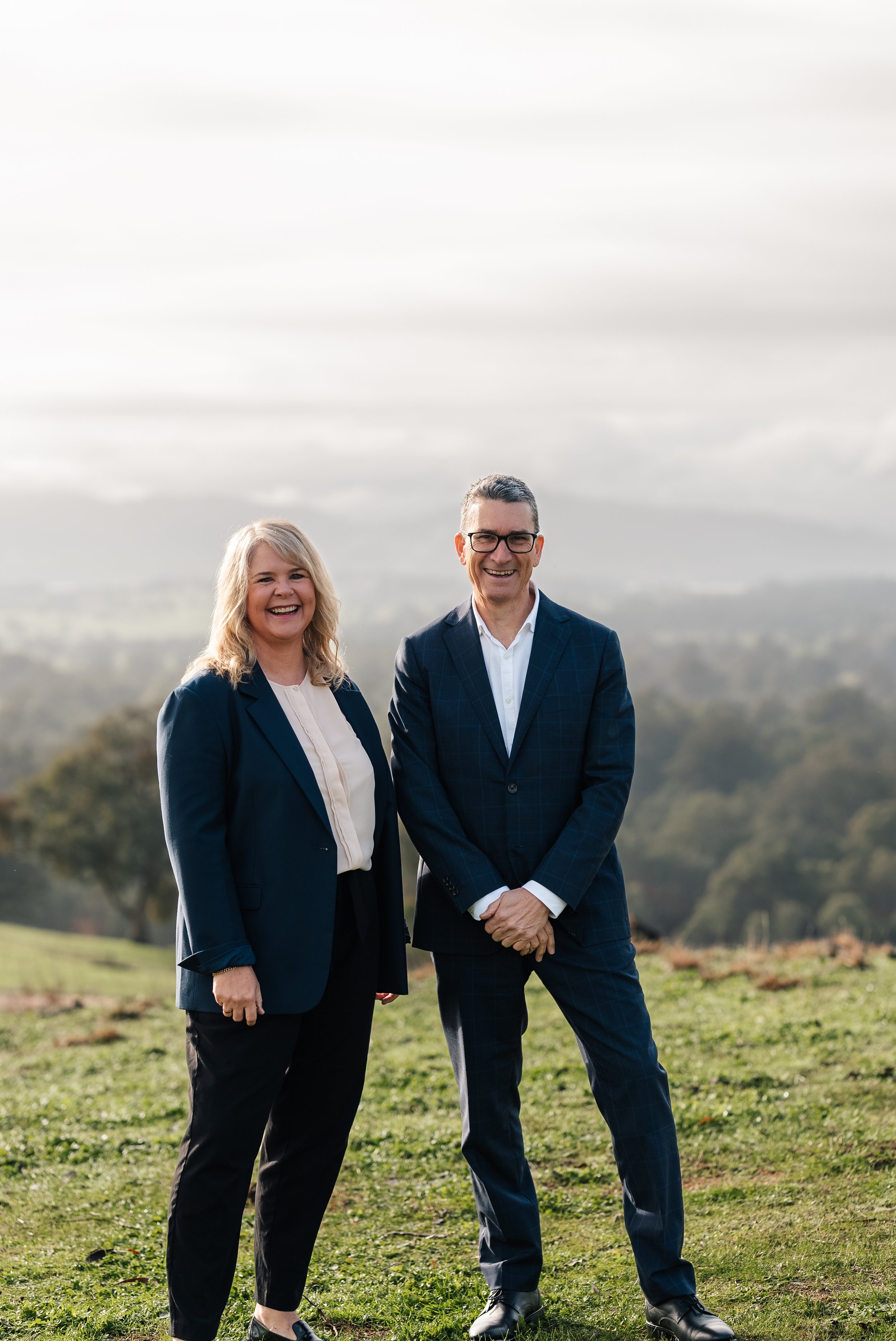 A man and a woman in business attire standing outdoors, with a scenic view of hills and trees in the background.