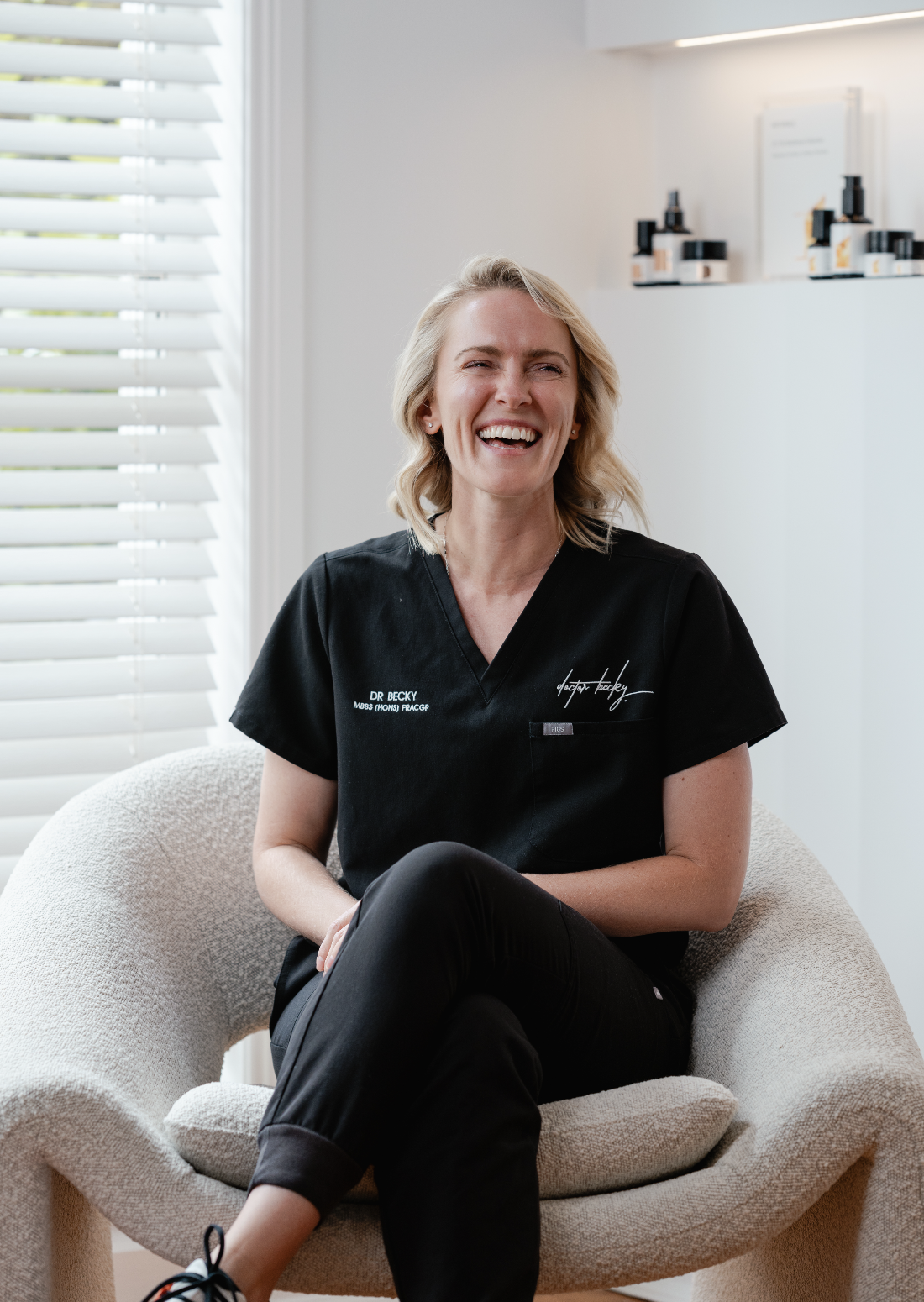 Smiling person in black medical scrubs sitting on a chair in a bright, modern room with white blinds and shelves with products in the background.