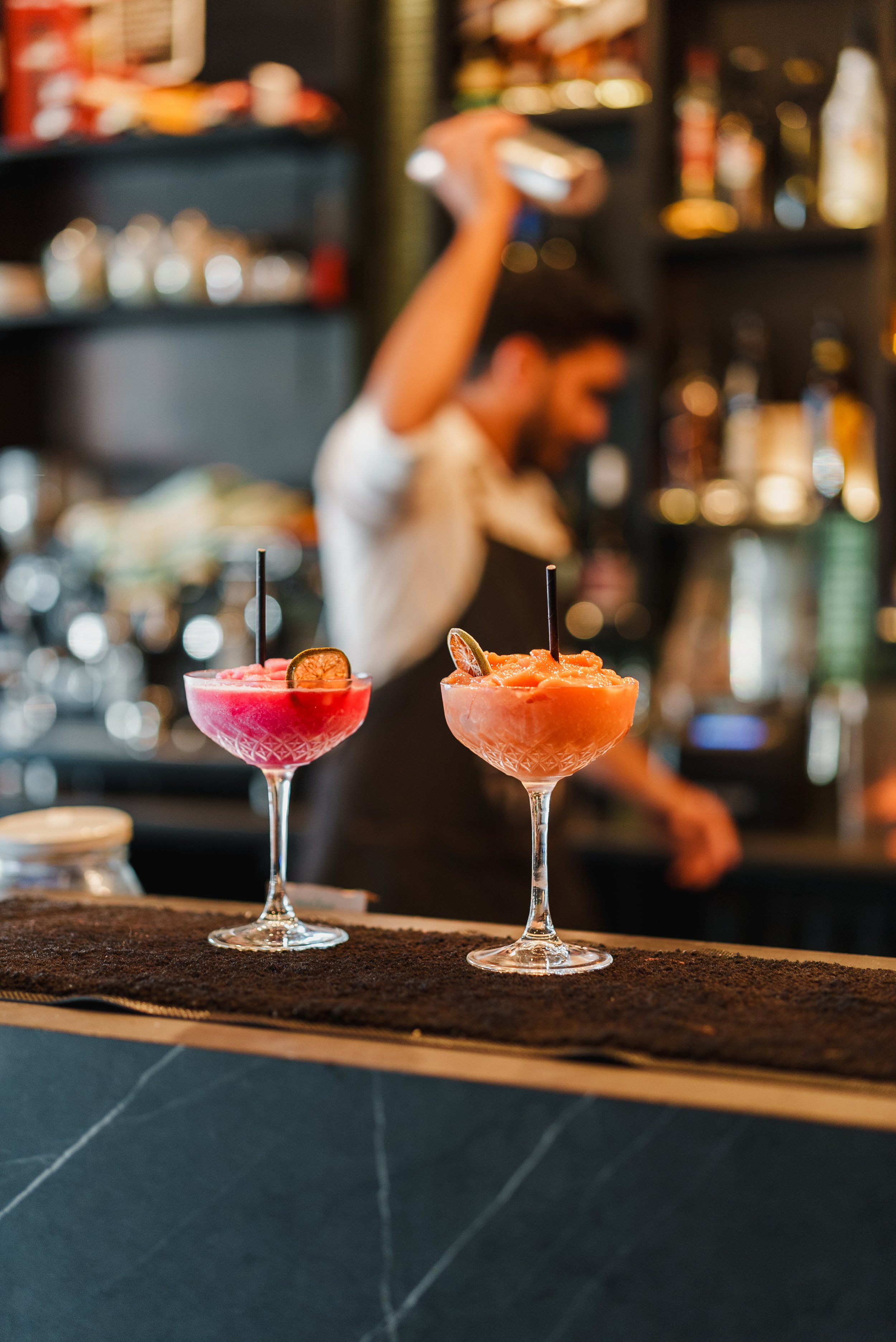 Two colorful cocktails in glasses with straws and garnishes on a bar counter, with a bartender shaking a cocktail shaker in the background.