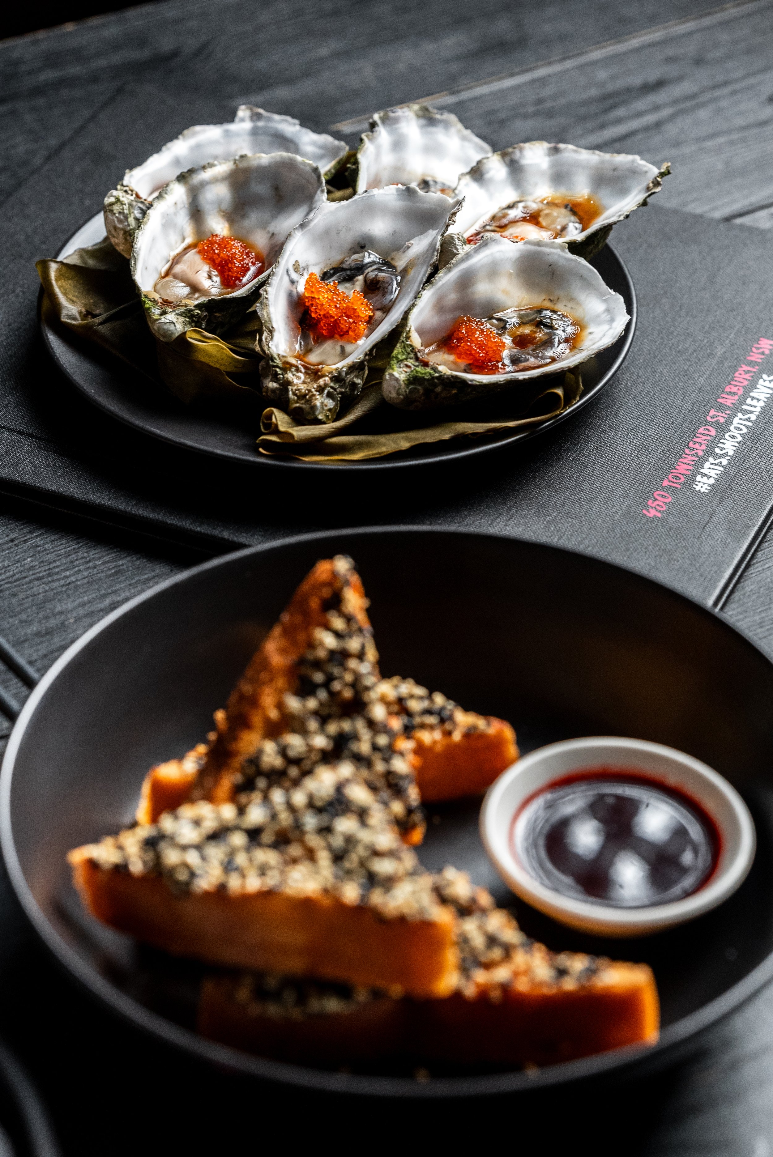 Gourmet dish with oysters topped with red caviar and Asian-inspired sesame-crusted toast with dipping sauce on plates.