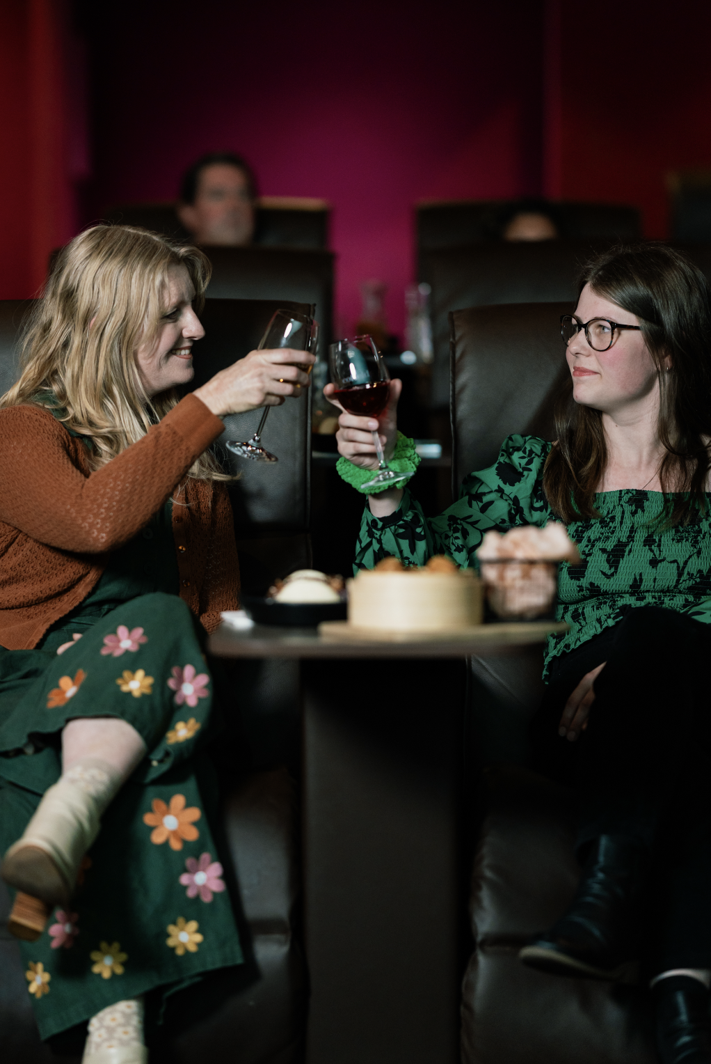 Two women sitting in a cozy lounge area, clinking wine glasses, with a small table holding food in front of them.