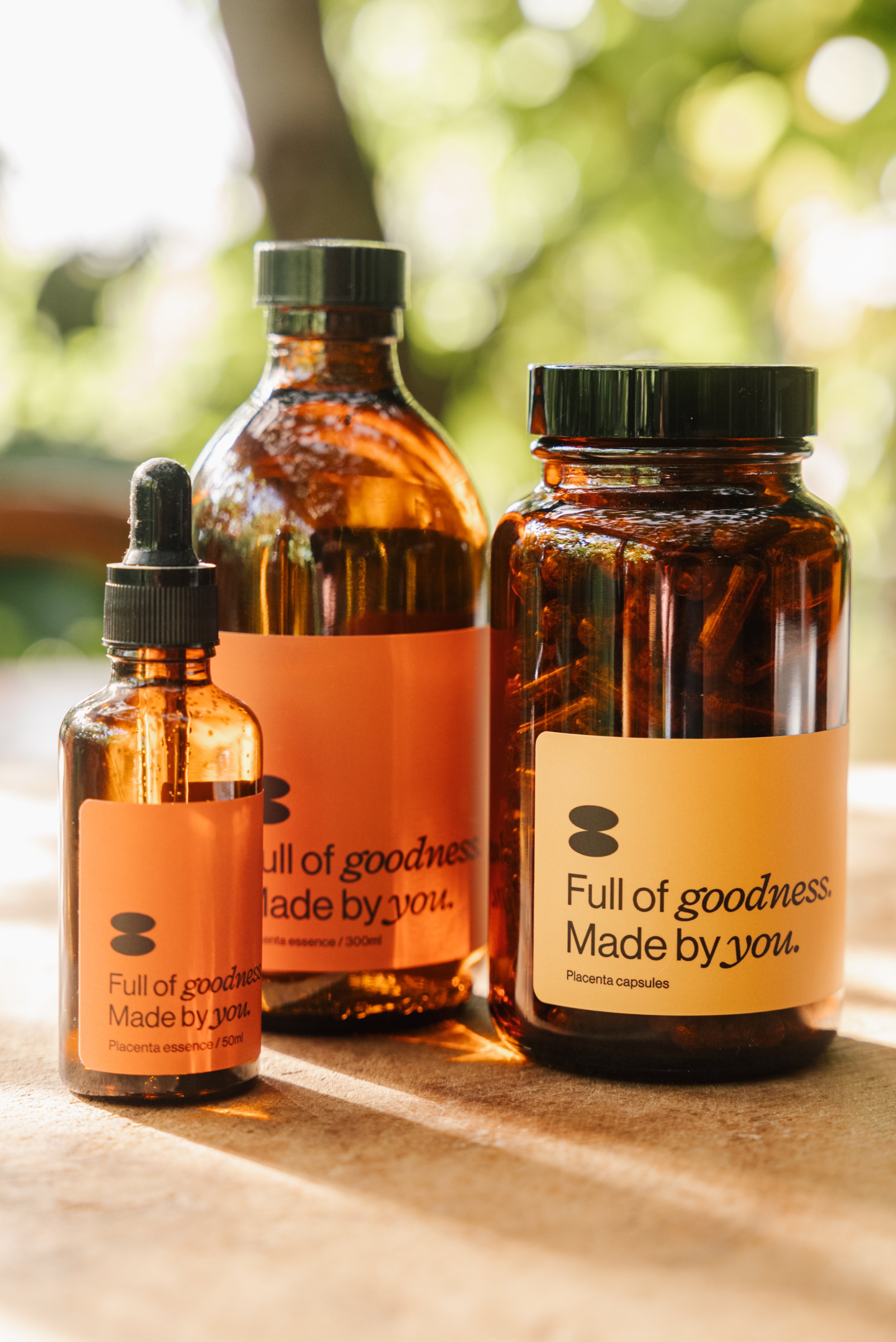 Three amber glass bottles with dropper and screw caps, labeled 'Full of goodness. Made by you. Placenta essence/capsules,' placed on a wooden surface against a blurry green background.