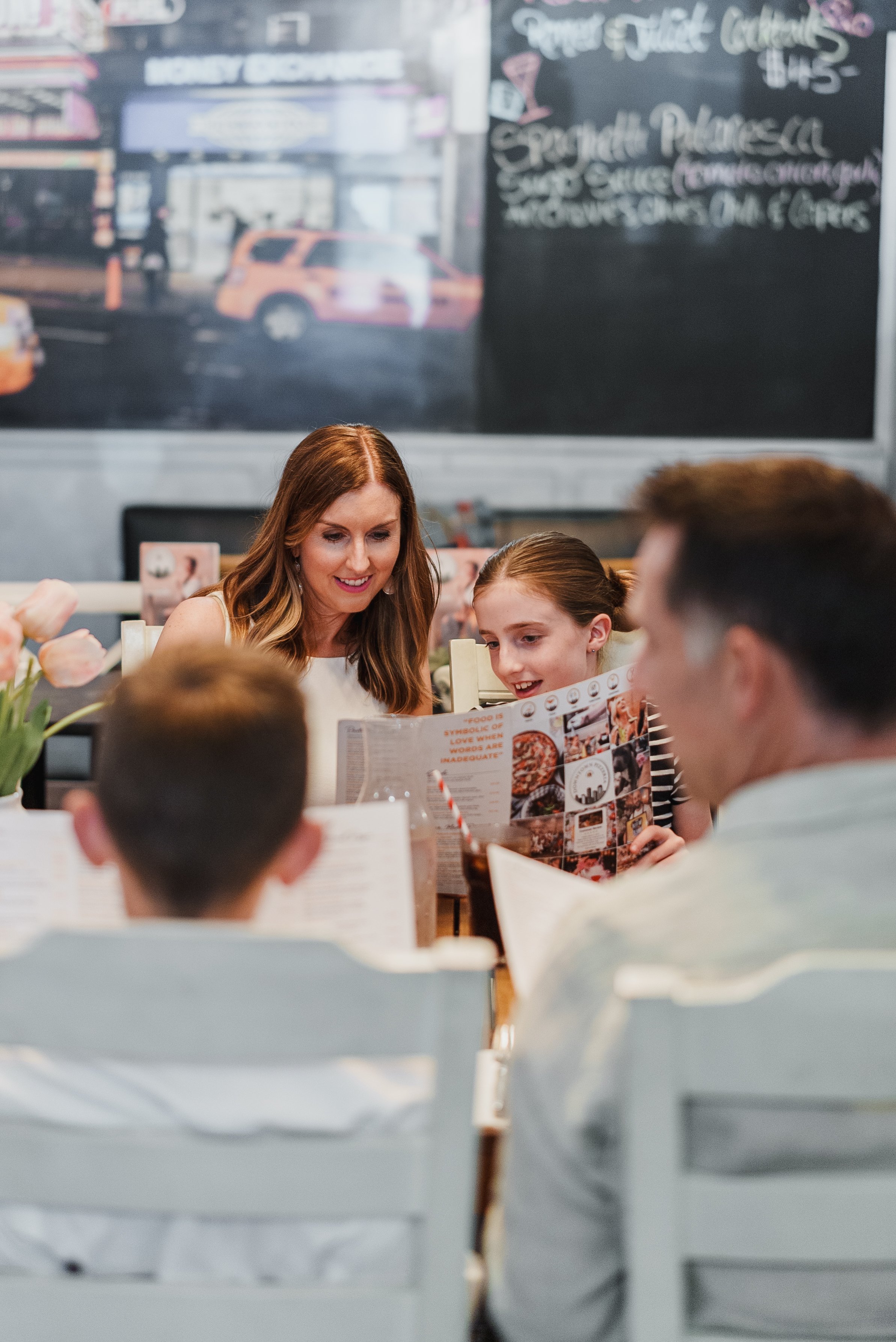 A family of four sitting at a restaurant table, looking at menus, with a chalkboard menu and street scene backdrop in the background.
