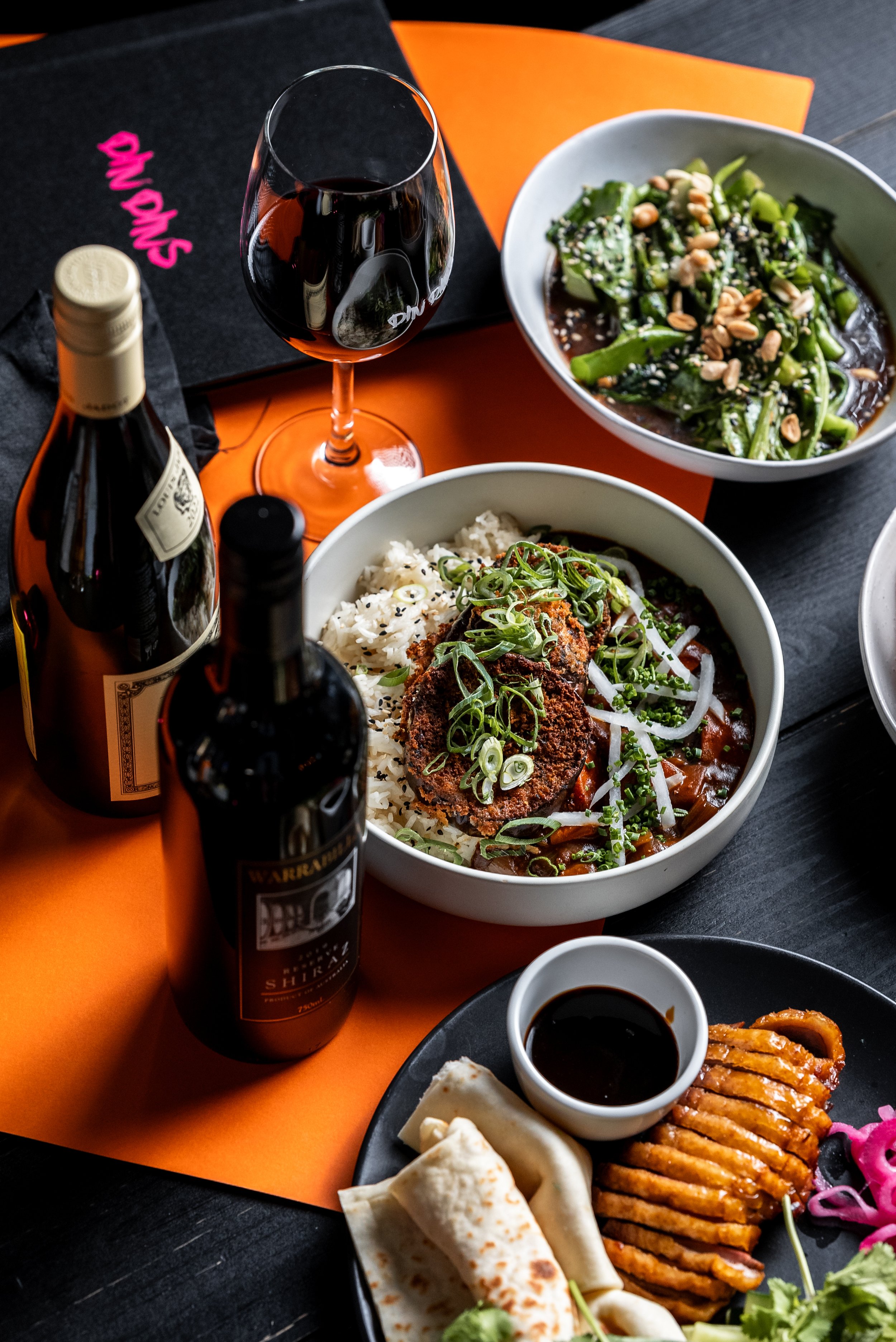 Assorted Asian dishes with wine; includes fried rice with cutlet, greens with peanuts, sliced duck, and sauce. Two wine bottles and a glass of red wine are present.