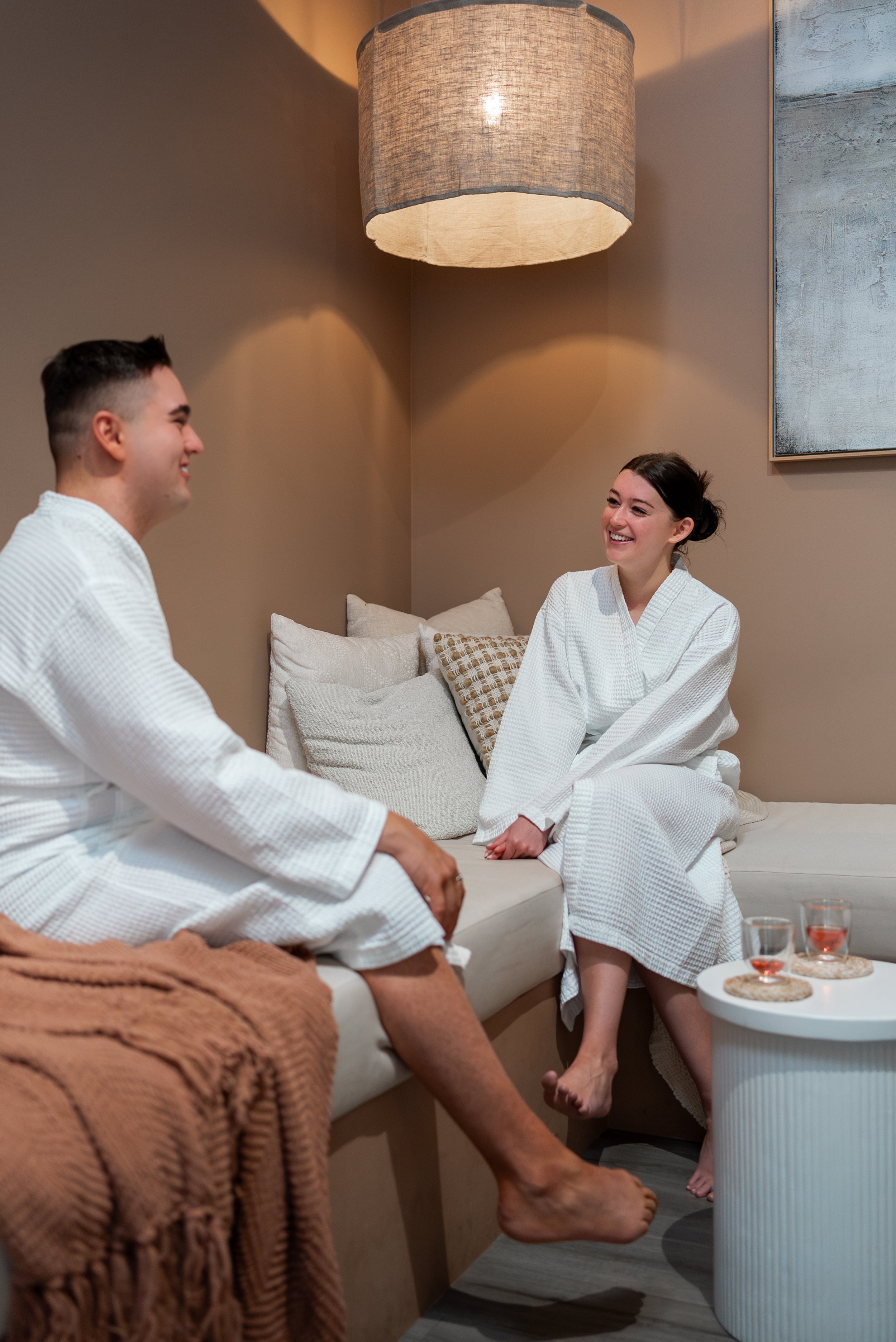 Two people in white bathrobes relaxing in a cozy lounge setting, sitting on a sofa with pillows. A hanging lamp above provides warm lighting. A small table with drinks is nearby, and a brown throw blanket is on the sofa.