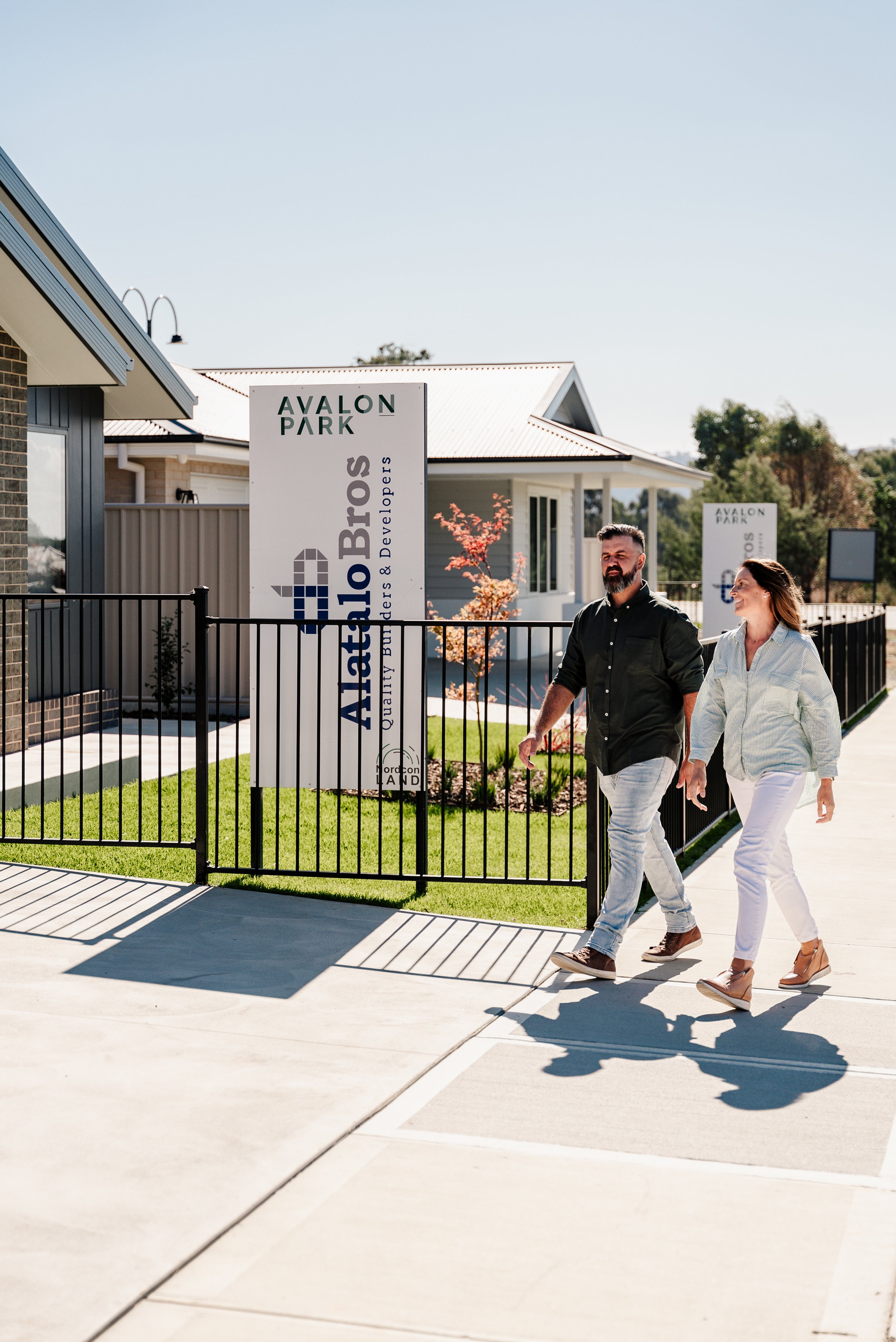 A couple walking on a sidewalk in front of a model home in Avalon Park, with a "Alatalo Bros" sign visible next to a fence, under a clear sky.