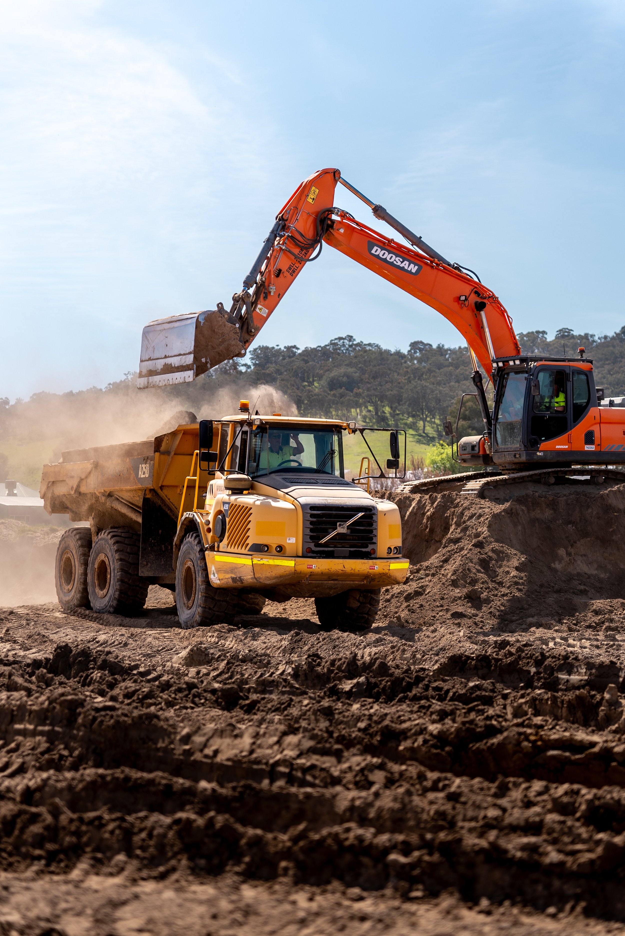 Excavator loading soil into a yellow dump truck on a construction site with trees in the background under a clear sky.
