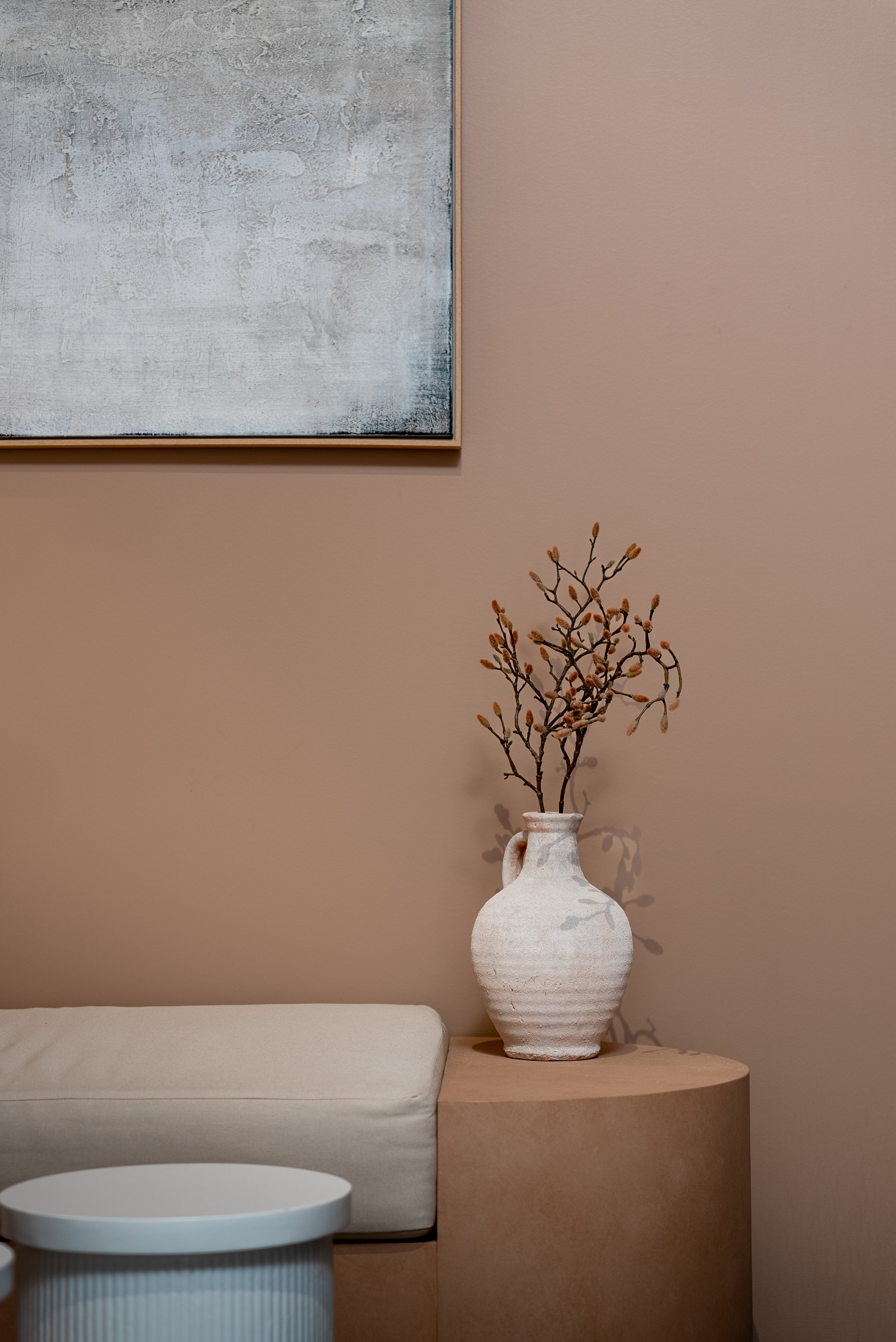 Minimalist interior with a beige wall, abstract painting, a rustic vase with dried branches, and beige furniture.