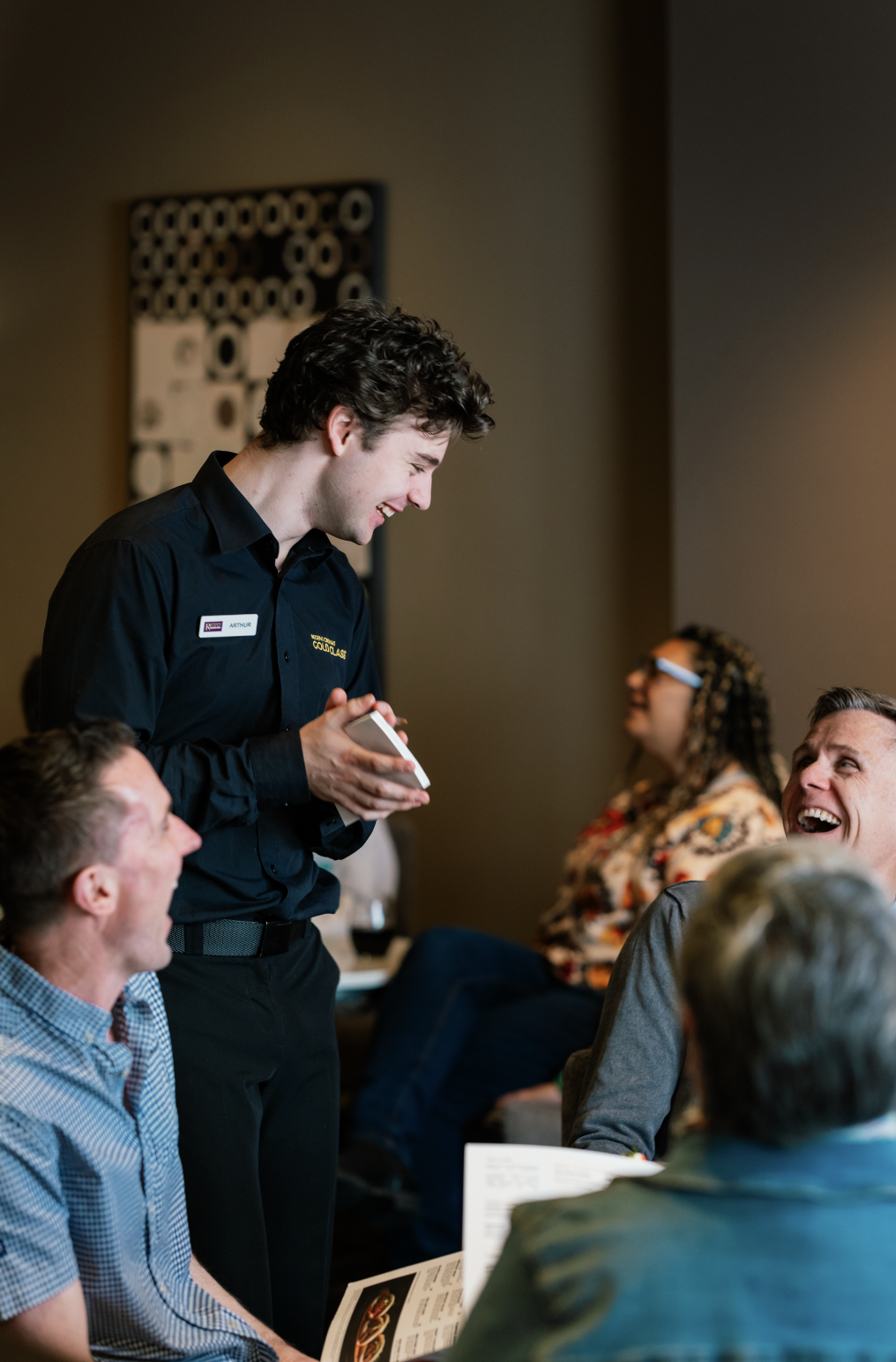 Waiter smiling and taking orders from a group of laughing customers in a restaurant.
