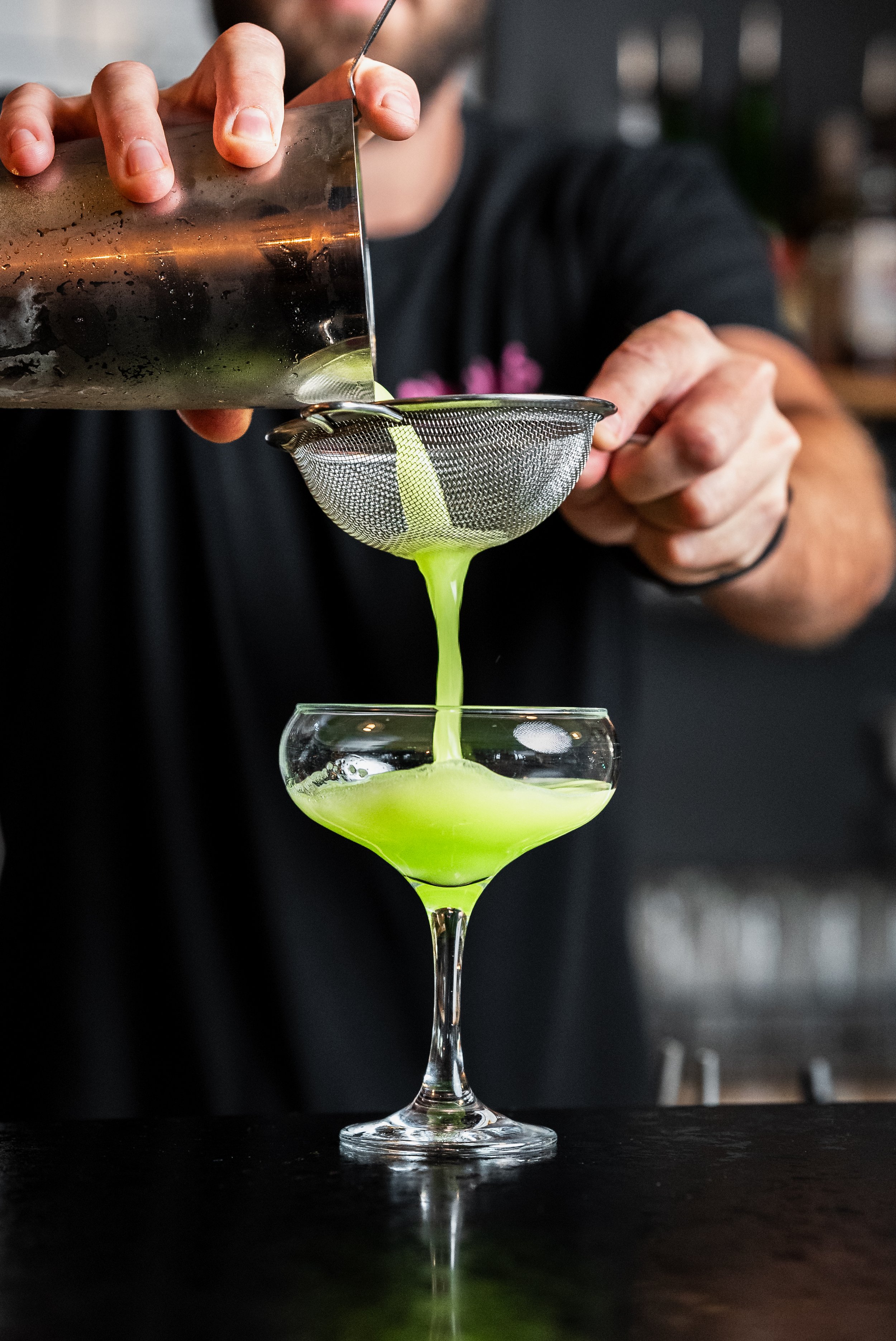Bartender pouring a green cocktail through a strainer into a coupe glass.