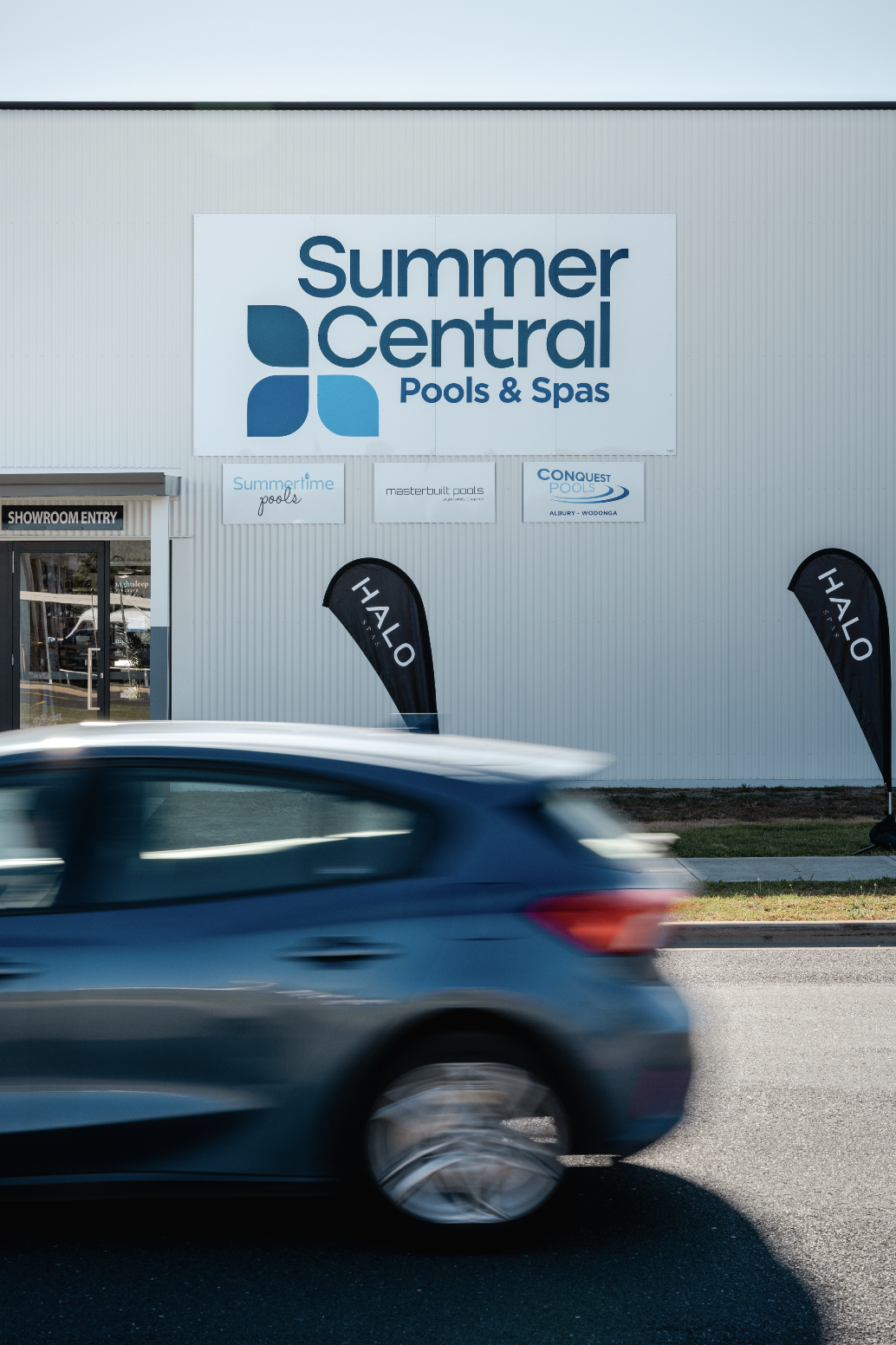 Exterior of Summer Central Pools & Spas building with signage, showroom entry, and a blue car passing by.