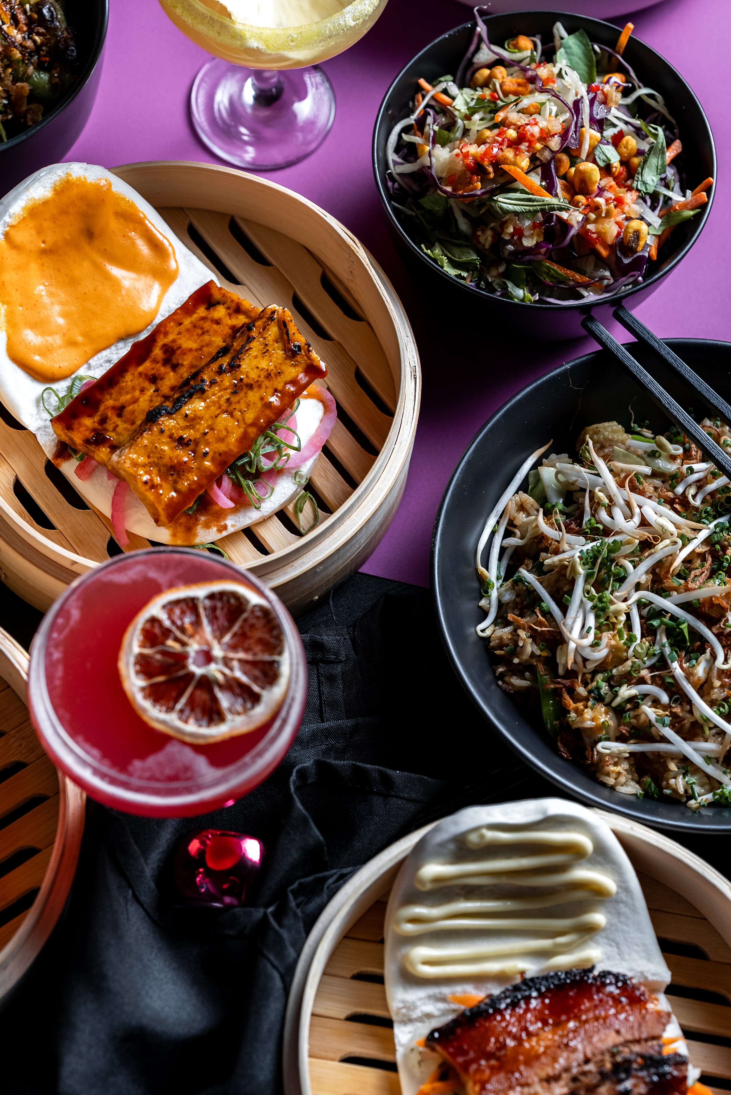 A table with an assortment of Asian-inspired dishes, including bao buns with grilled meat, a bowl of salad with peanuts and vegetables, a noodle dish with bean sprouts, and two cocktails, one with a dried citrus garnish, on a purple surface.
