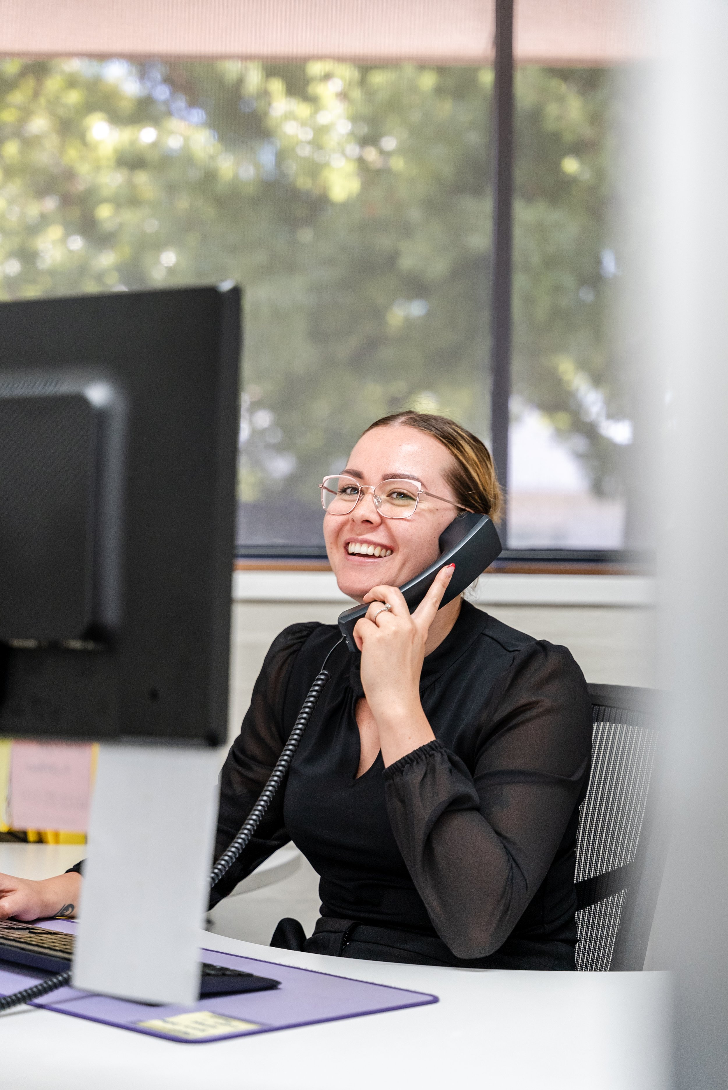 Woman in an office talking on a landline phone while using a computer, wearing glasses and a black blouse.