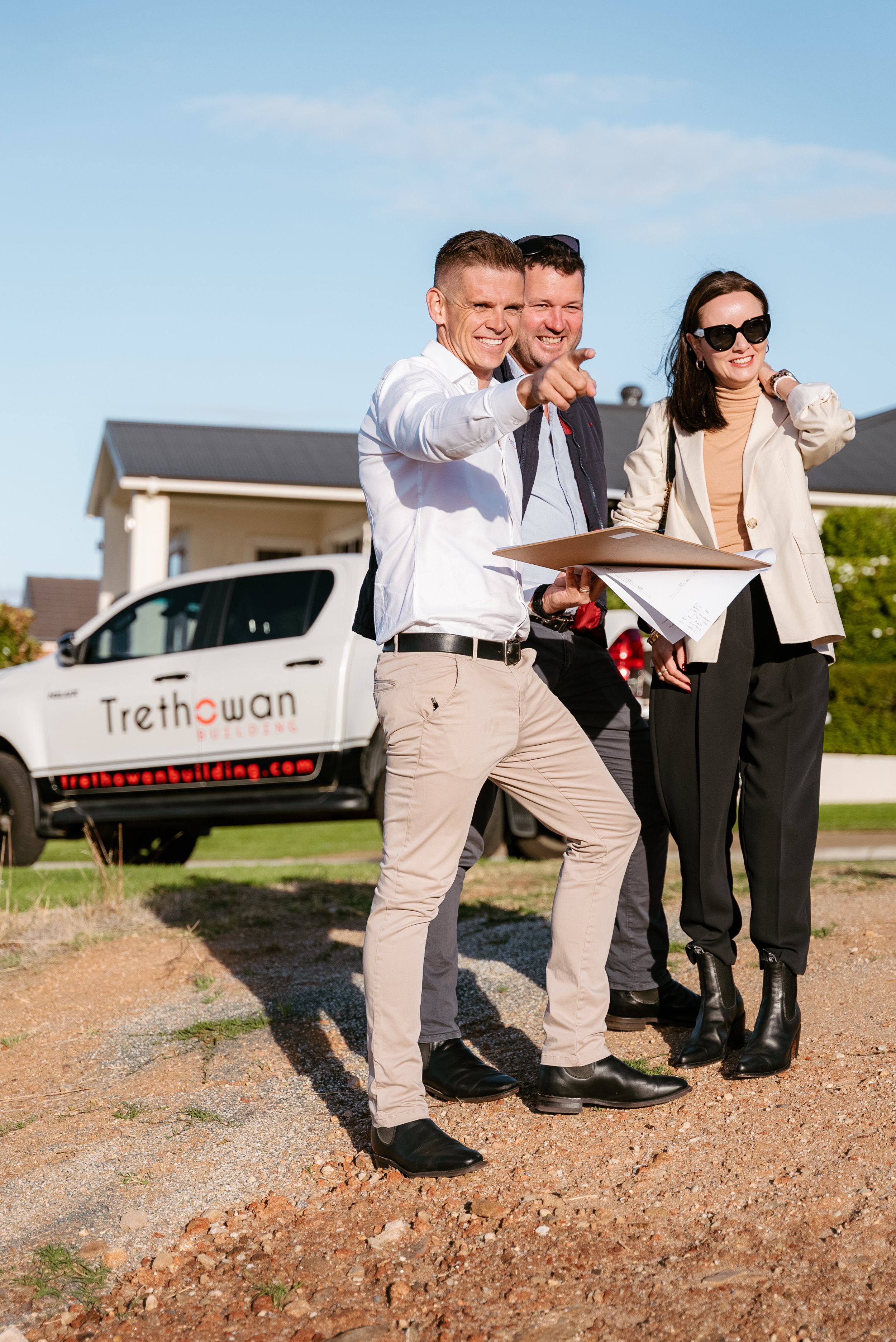 Three people outside on a construction site, two men and a woman, discussing plans. The woman holds a folder, and a vehicle with 'Trethowan Building' branding is in the background.