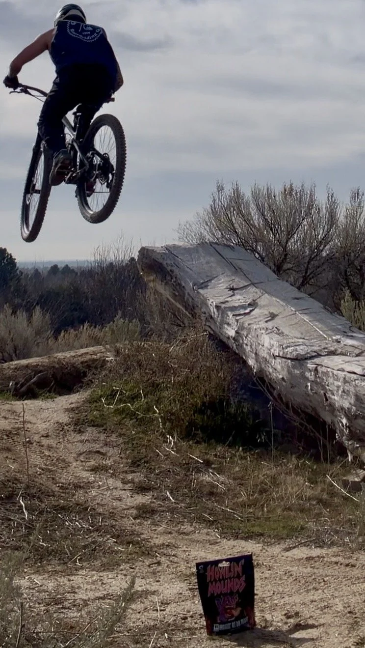 A person riding a mountain bike in mid-air over a dirt trail and a fallen log during daytime under an overcast sky with a bag of send it snacks almonds laying on the ground.