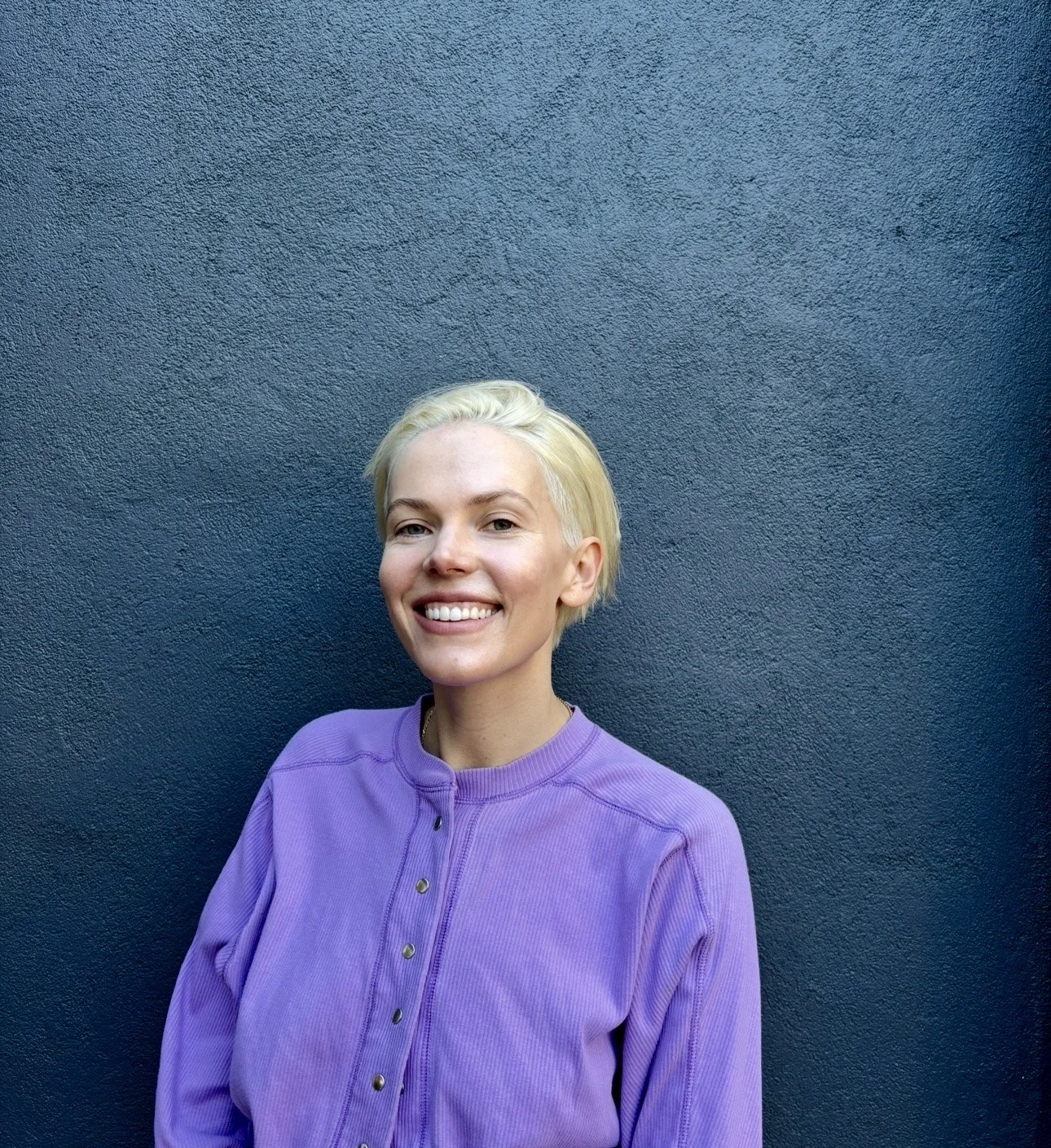 A woman with short blonde hair smiling against a dark blue textured wall, wearing a purple long-sleeve shirt.