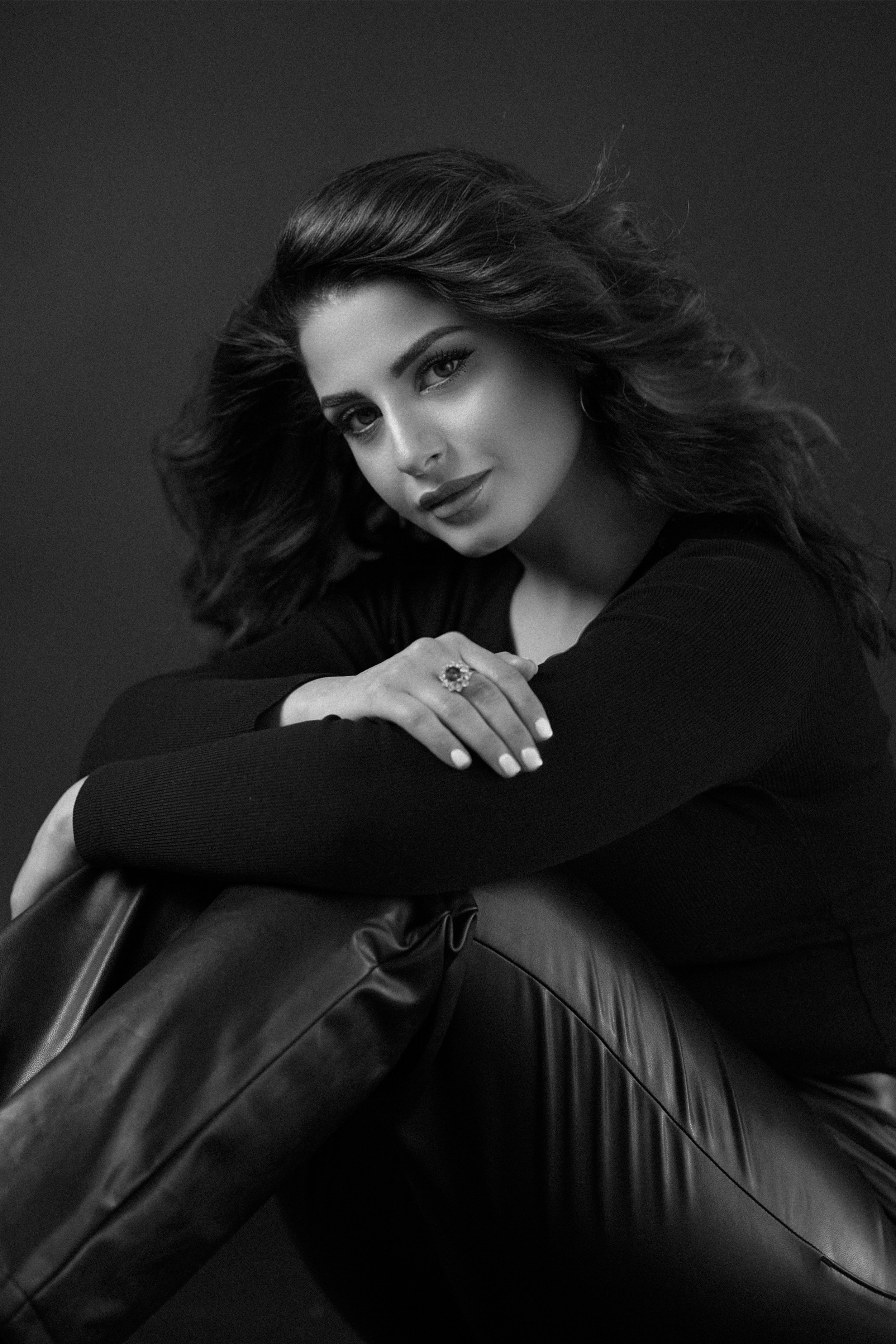 Black and white portrait of a woman with wavy hair, sitting and looking at the camera, wearing a dark long-sleeve top and leather pants.