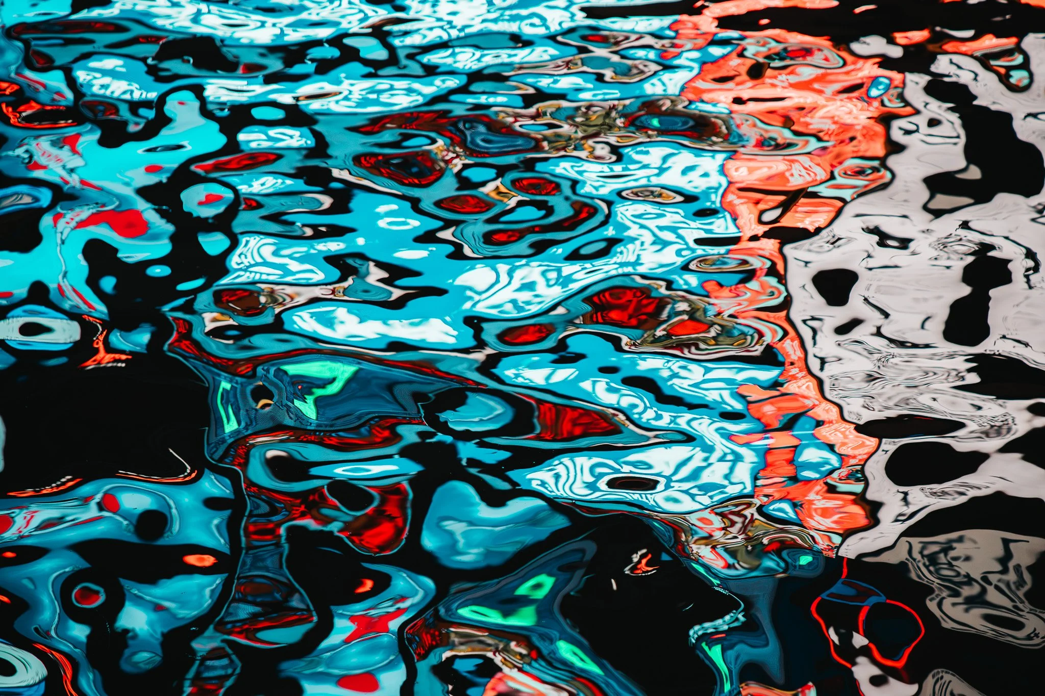 Abstract view of water with colorful reflections including blue, red, white, black, and some green hues, showing ripples and distortions.