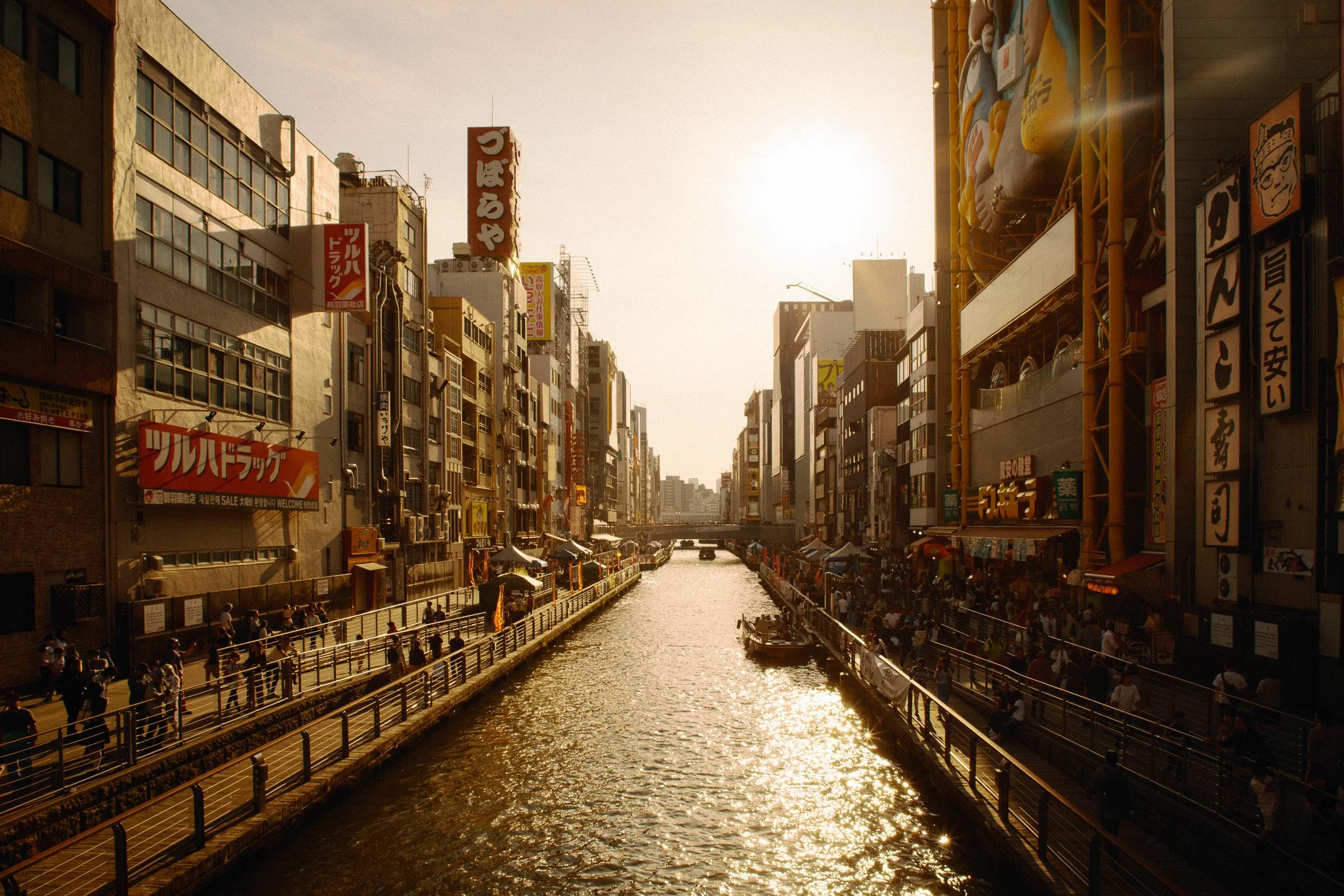 City canal during sunset with buildings and pedestrians on both sides, boats on the water, and various signs in Japanese.