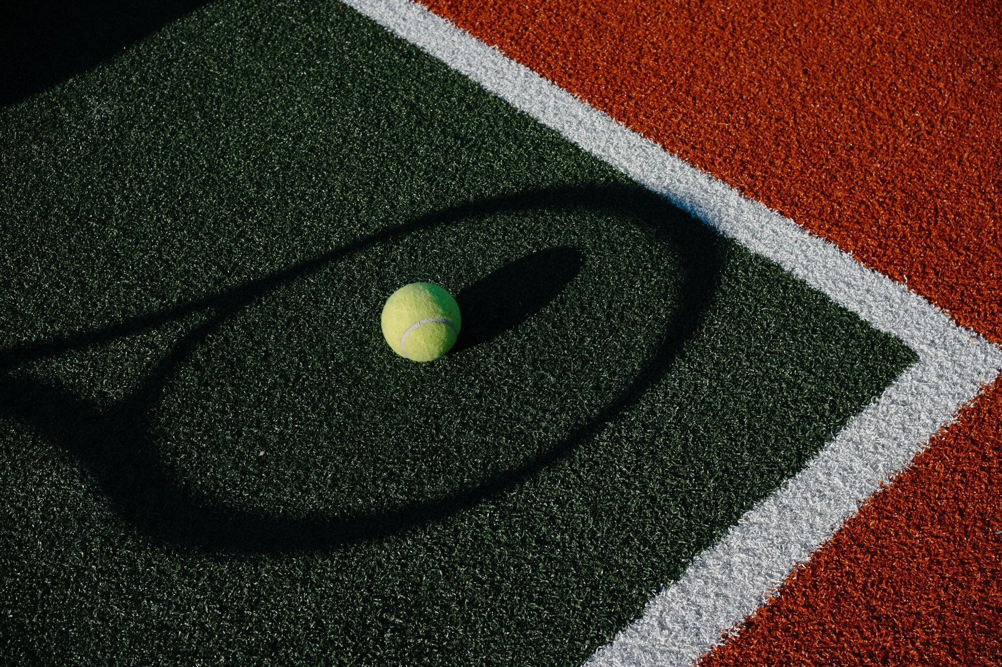A green tennis ball lies on a tennis court with green, white, and red textured surfaces. The ball casts a shadow on the court.