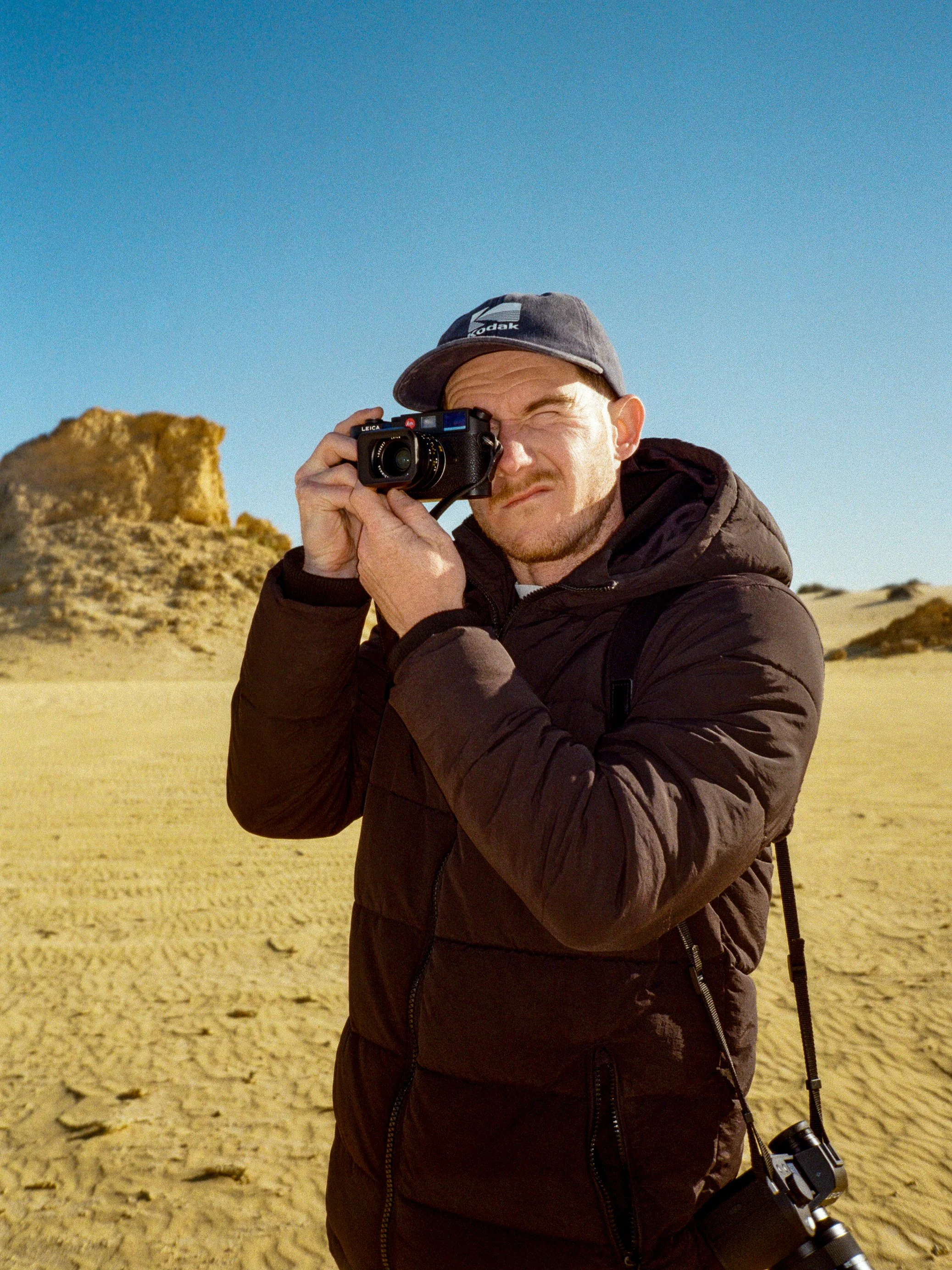 A man dressed in a brown jacket and cap is aiming a camera in a desert environment with sandy terrain and rock formations under a clear blue sky.