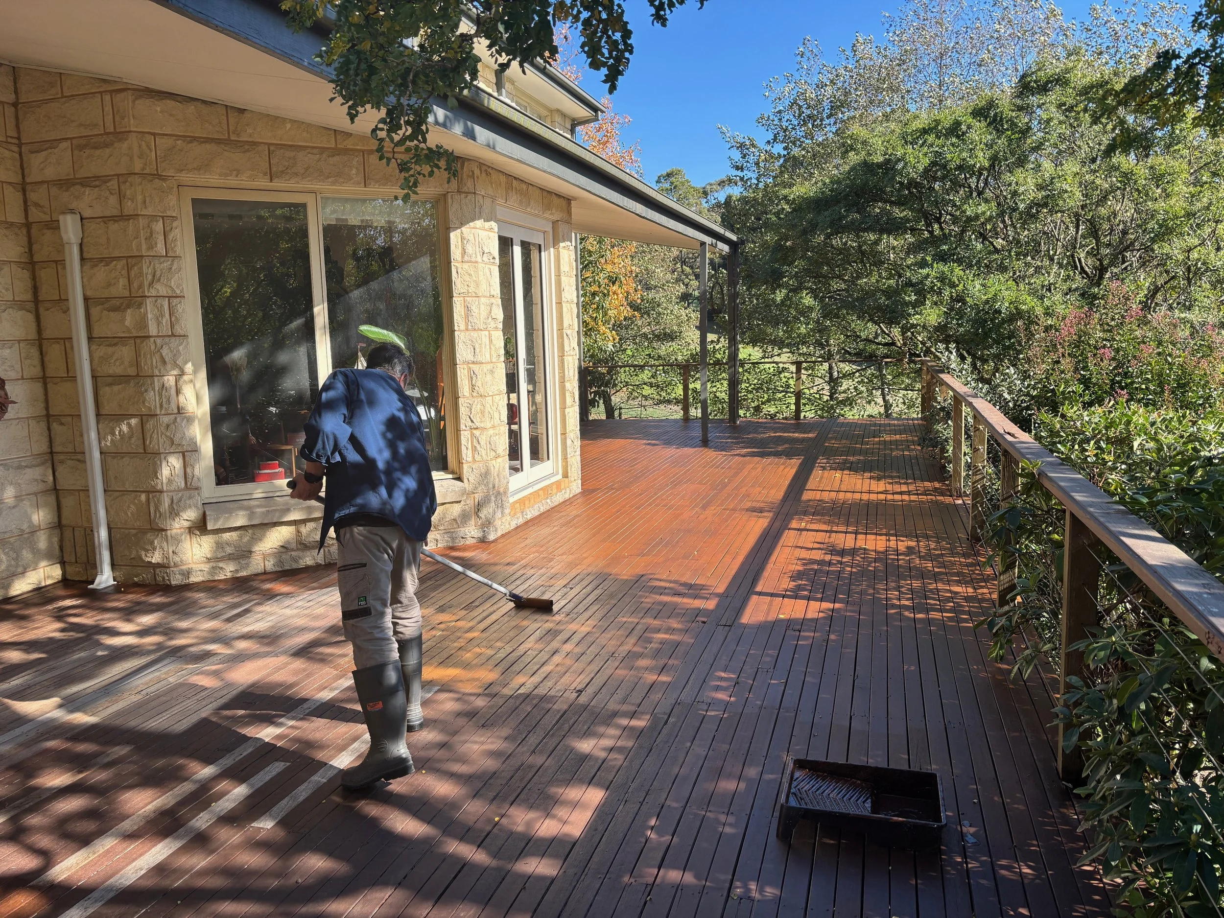 Person wearing rubber boots and navy jacket cleaning a wooden deck with a broom on a sunny day, beside a house with large glass windows, surrounded by lush green trees.