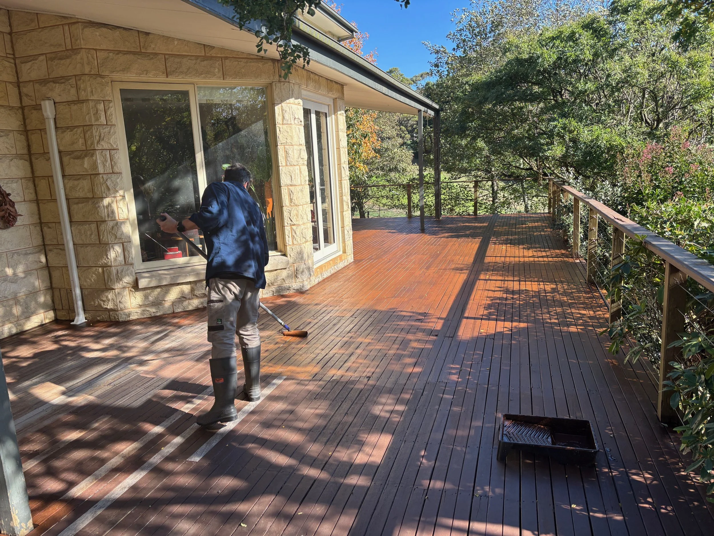 A person cleaning a wooden deck outside a house on a sunny day, with trees and sky visible in the background.