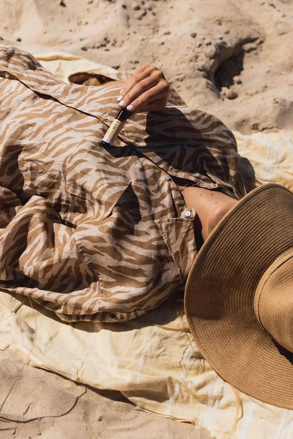 Person lying on the beach wearing a zebra-patterned shirt, holding a small bottle, with a wide-brimmed straw hat next to them.