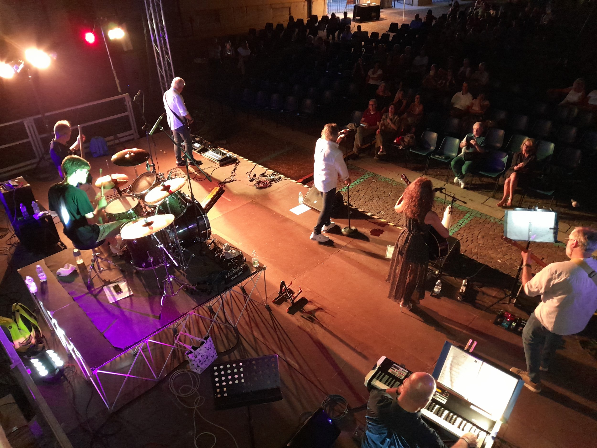 I Nuovi solidi che si esibiscono su un palco con strumenti dal vivo, mentre un pubblico di spettatori è seduto in un'area di ascolto all'aperto, in serata