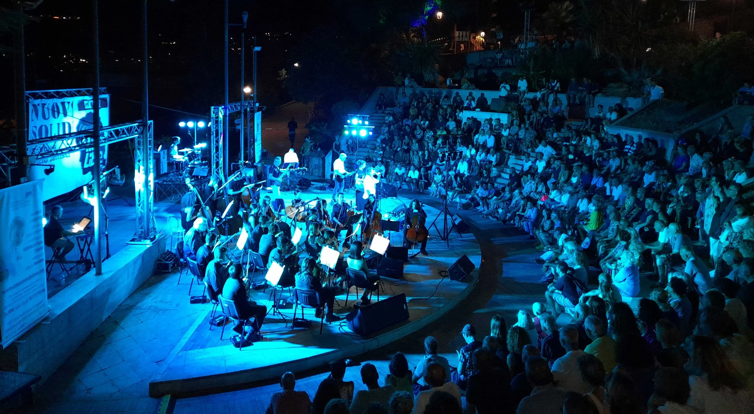 Concerto all'aperto con orchestra in abiti scuri, illuminato in blu, davanti a un pubblico seduto in gradinate, di notte