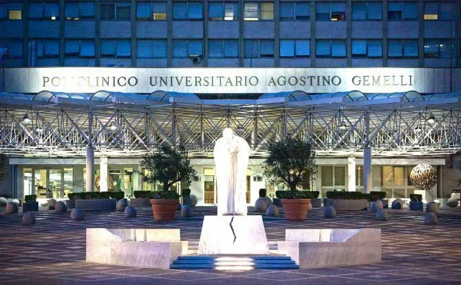 Entrata del Policlinico Universitario Agostino Gemelli, con statua in primo piano, piante e luci decorative.