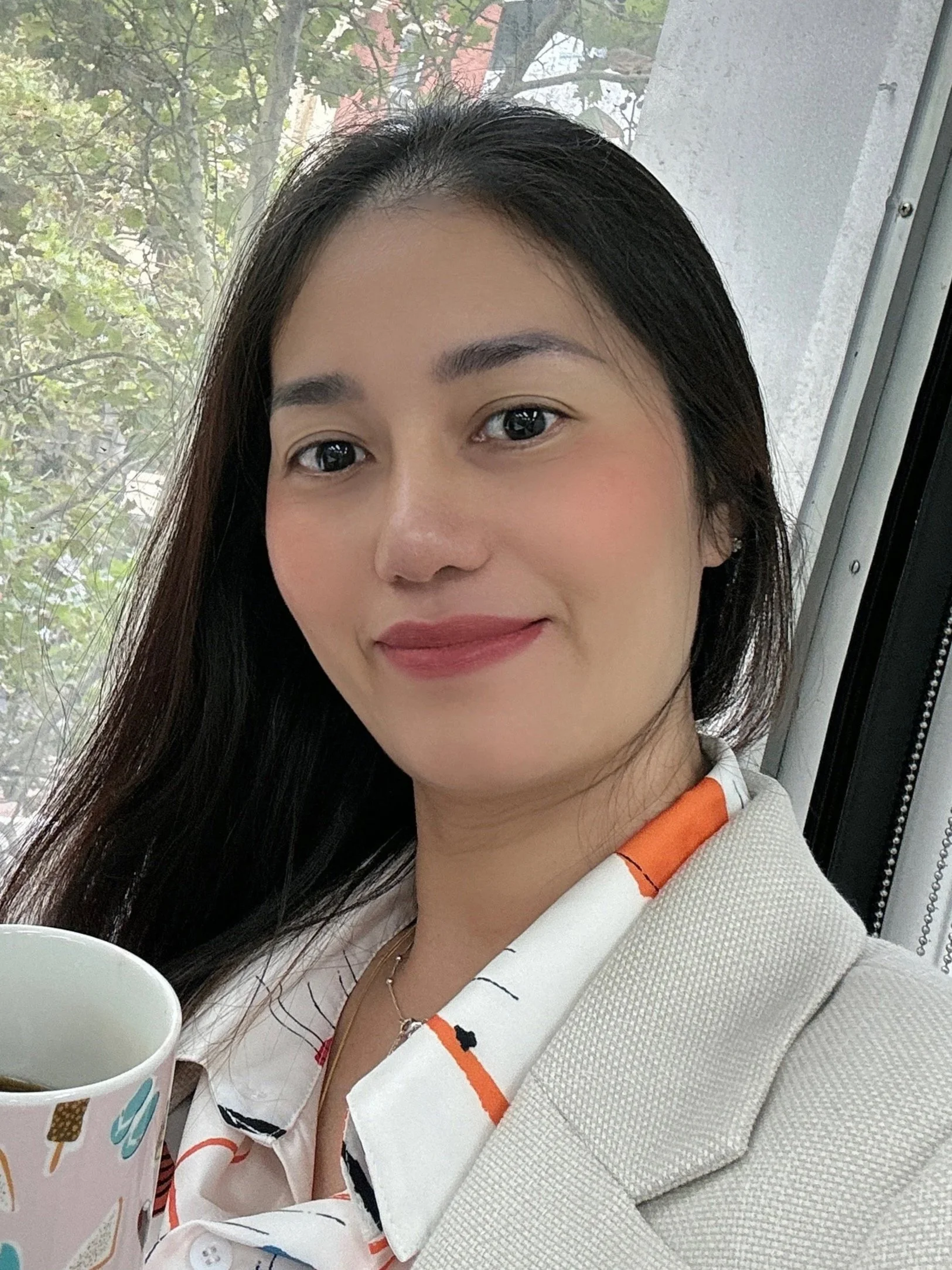Close-up of a smiling woman with curly hair, wearing glasses, hoop earrings, layered necklaces, and a striped blouse indoors.