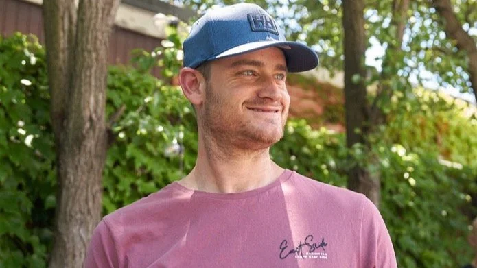 A man smiling outdoors wearing a blue baseball cap and a pink t-shirt with a signature on it.