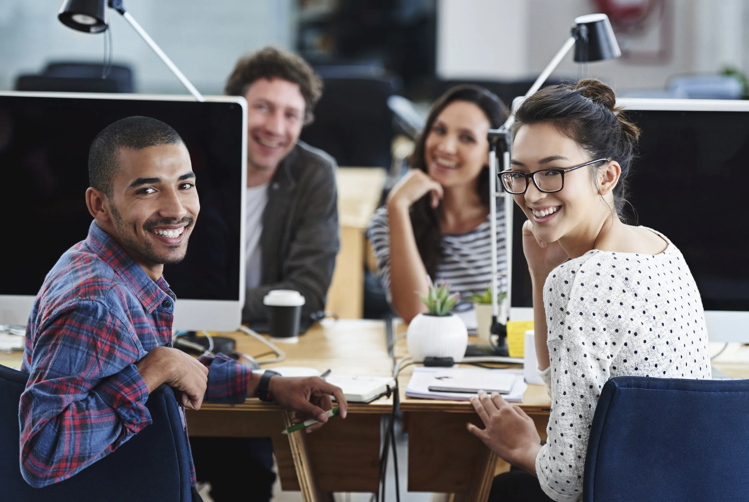 Vier Personen sitzen um einen Tisch in einem Büro, lachen und sprechen. Im Vordergrund links sitzt ein Mann in einem karierten Hemd, im Vordergrund rechts eine Frau mit Brille und Polka Dot-Shirt. In der Hintergrundmitte zwei weitere Personen, ein Mann und eine Frau, die beide lächeln.