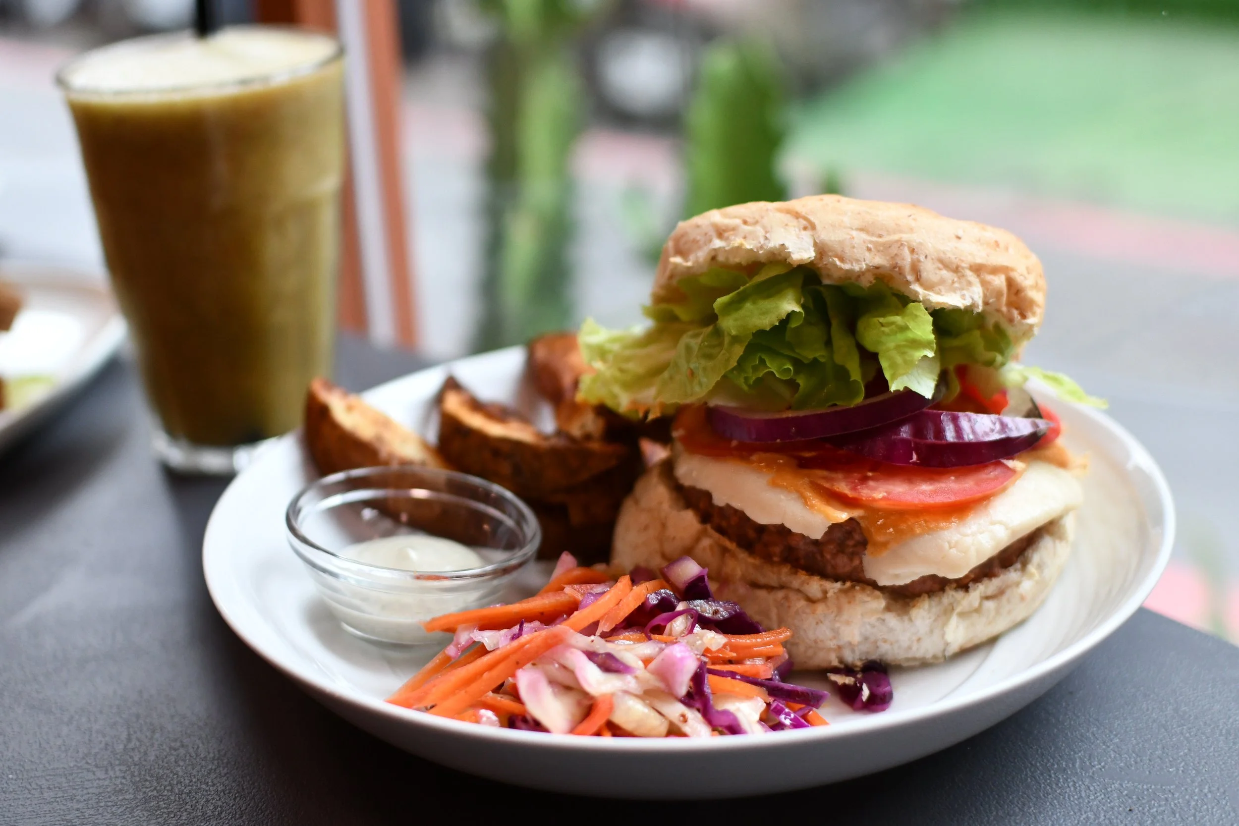 A plate with a burger topped with lettuce, tomato, onions, and cheese, served with potato wedges, coleslaw, and a small cup of dipping sauce. In the background, there is a glass of green smoothie or juice.
