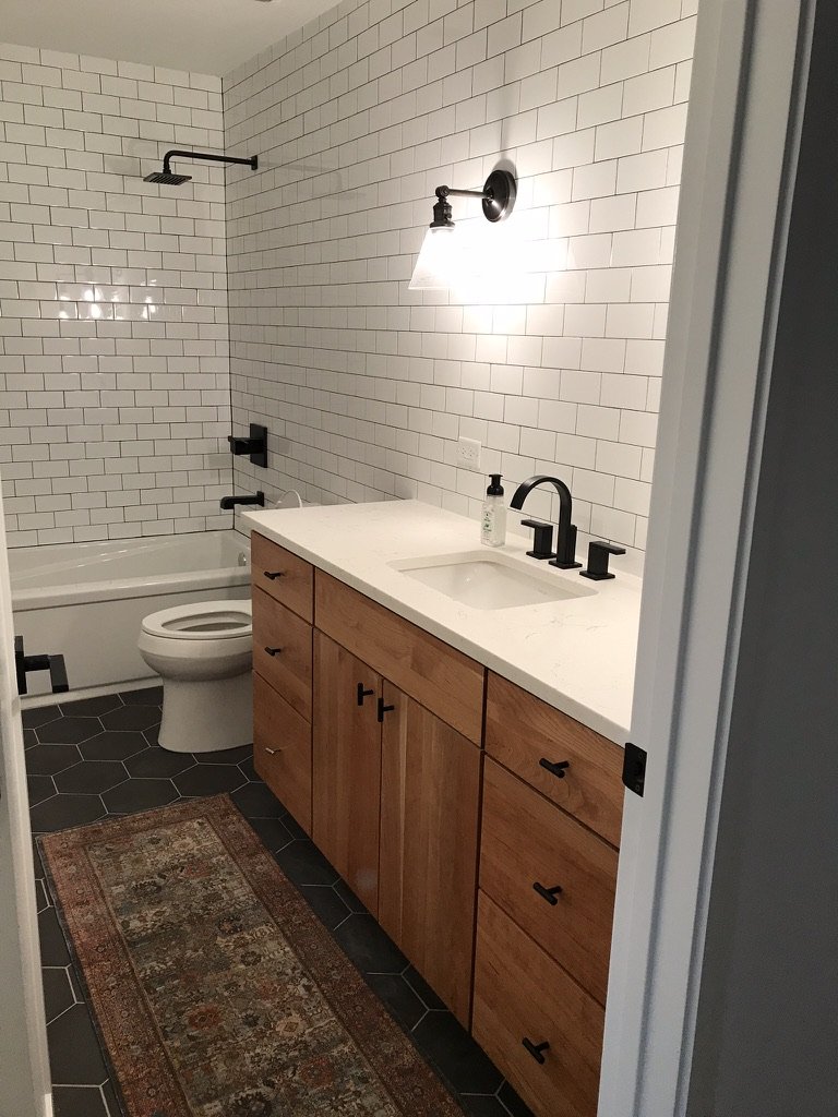 Modern bathroom with white subway tiles, wooden vanity with white countertop, black fixtures, hexagonal dark floor tiles, and a patterned rug.