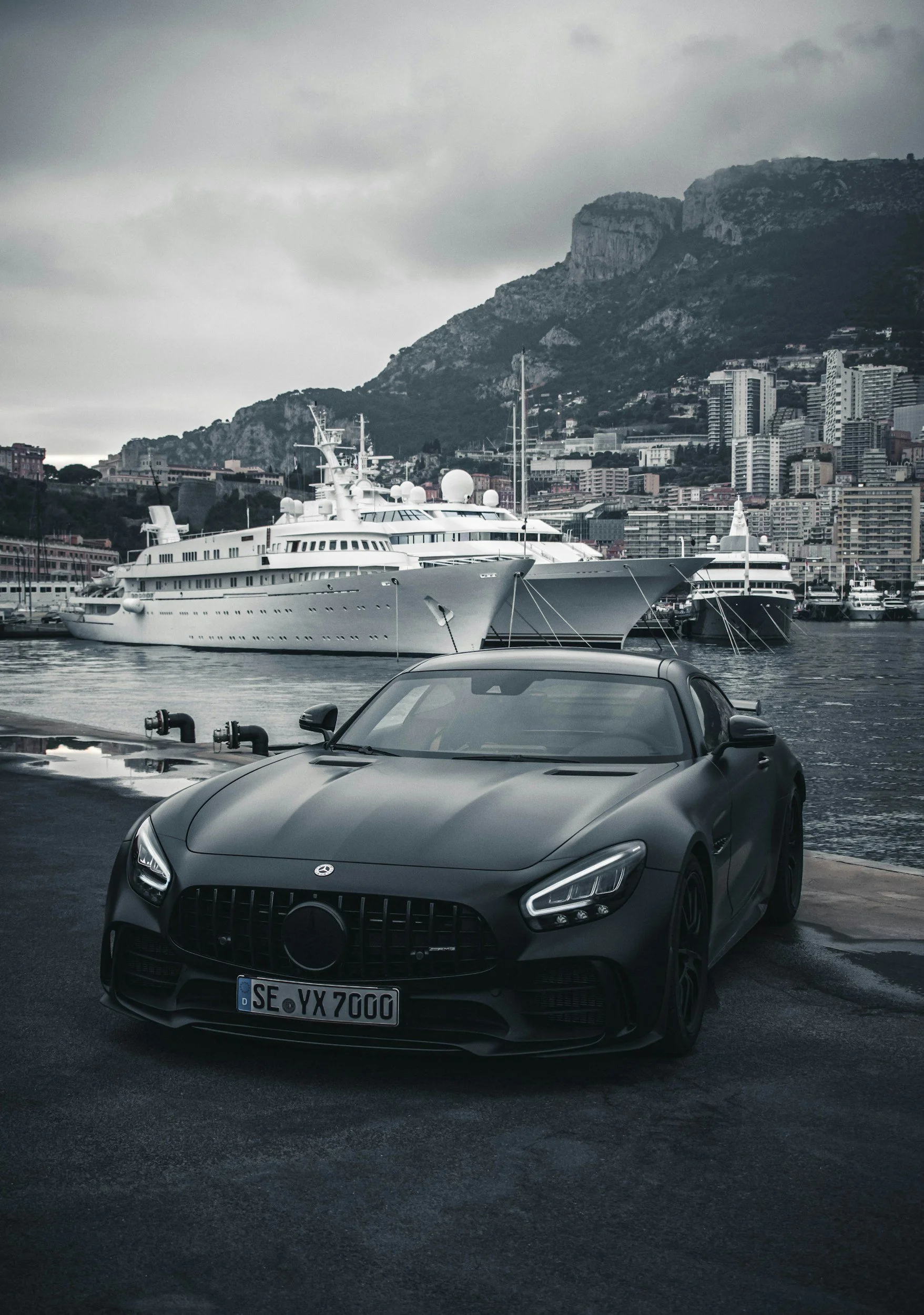 Voiture de sport noire garée au bord de l'eau avec des yachts en arrière-plan et une ville construite en hauteur sous un ciel nuageux.