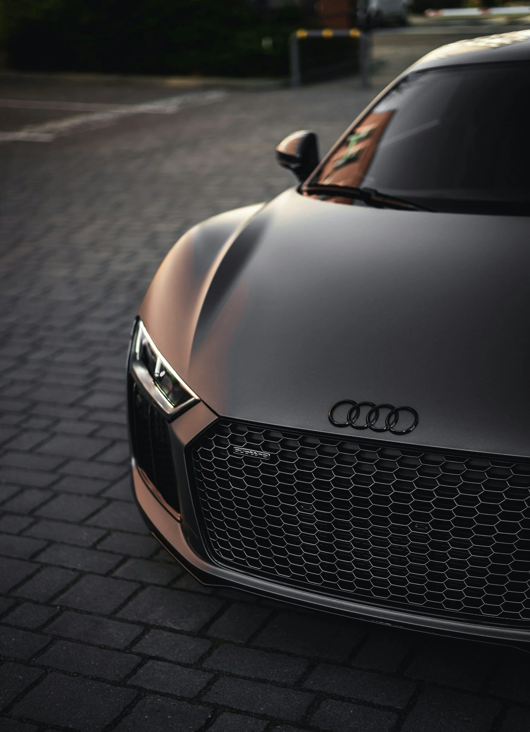 Première moitié avant d'une voiture noire Audi sport avec un logo sur le capot et une grille de radiateur en forme de nid d'abeille, stationnée sur un pavé, en soirée ou au crépuscule.