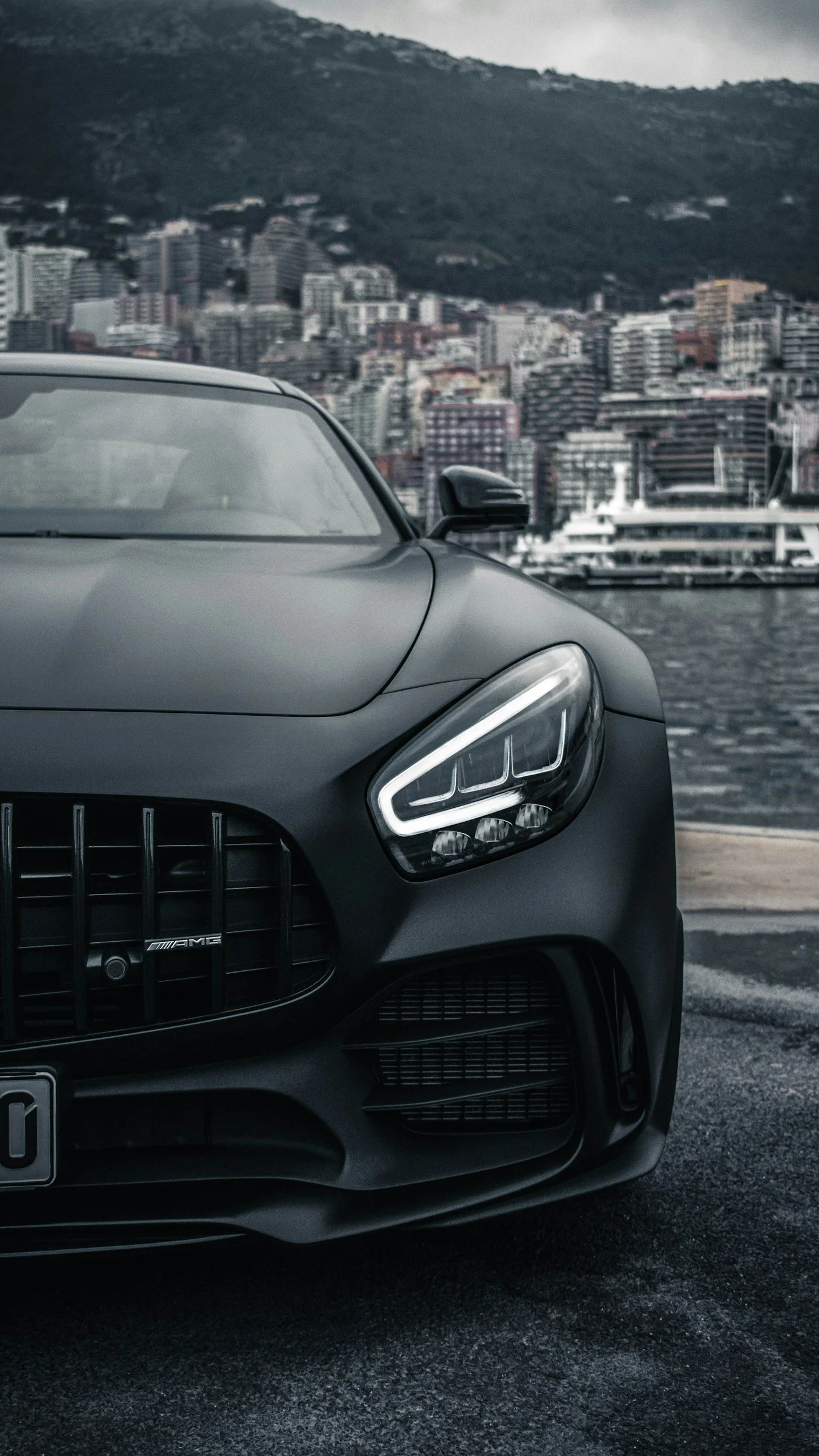 Une voiture noire de luxe, probablement une Mercedes AMG, garée près d'un port avec une ville en arrière-plan, comprenant des immeubles et une montagne.