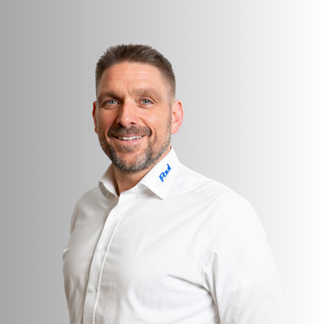 Portrait eines lachenden Mannes mit grauem Hintergrund, trägt weißes Hemd mit 'Paul' am Kragen.
