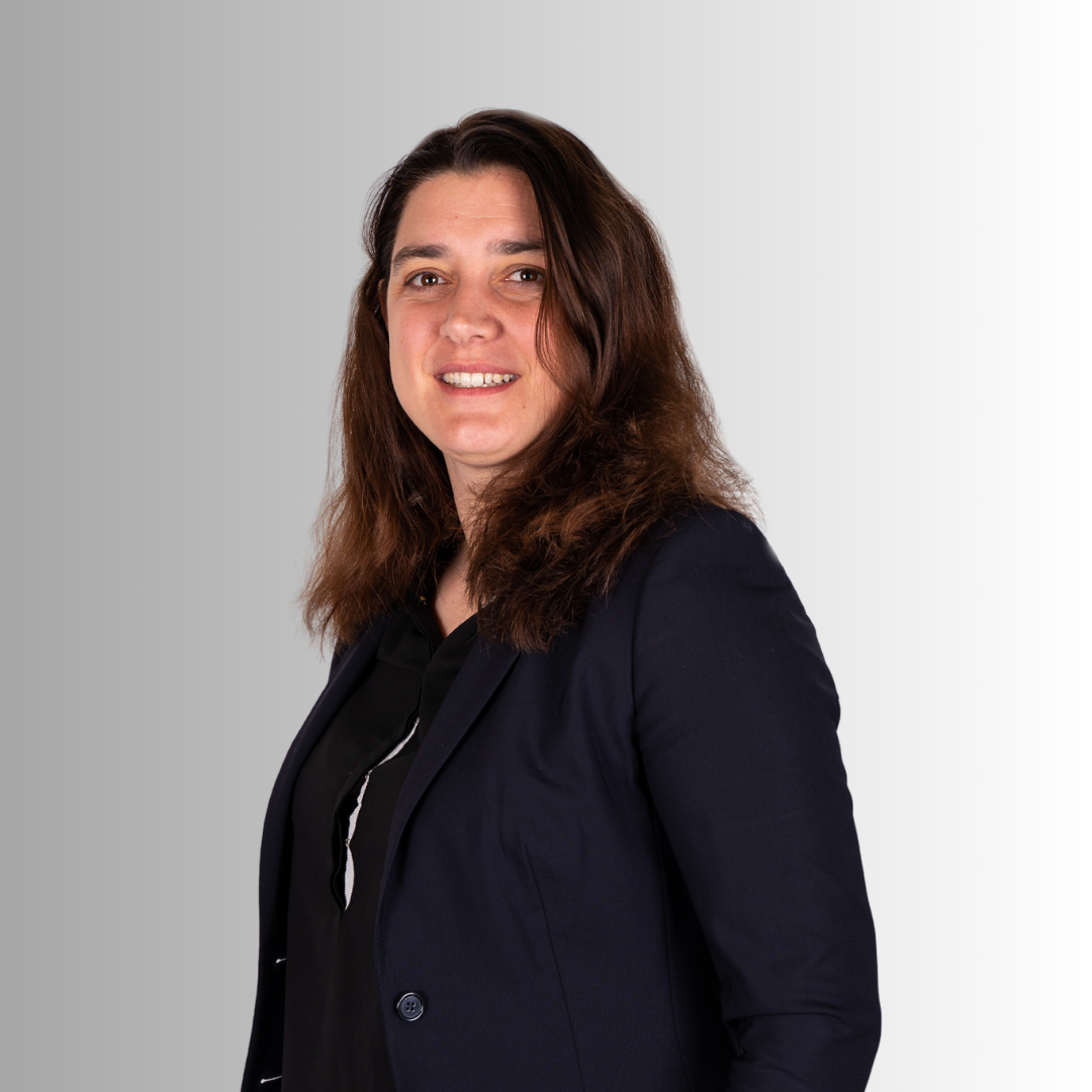 Porträt einer lächelnden Frau mit braunen Haaren, dunklem Blazer und schwarzem Hemd vor grauem und weißem Hintergrund.