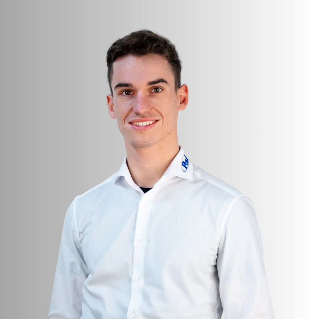 Junger Mann mit dunklen Haaren trägt weißen Hemd mit blauer AufSchrift am Kragen, lächelt vor grauem Hintergrund.