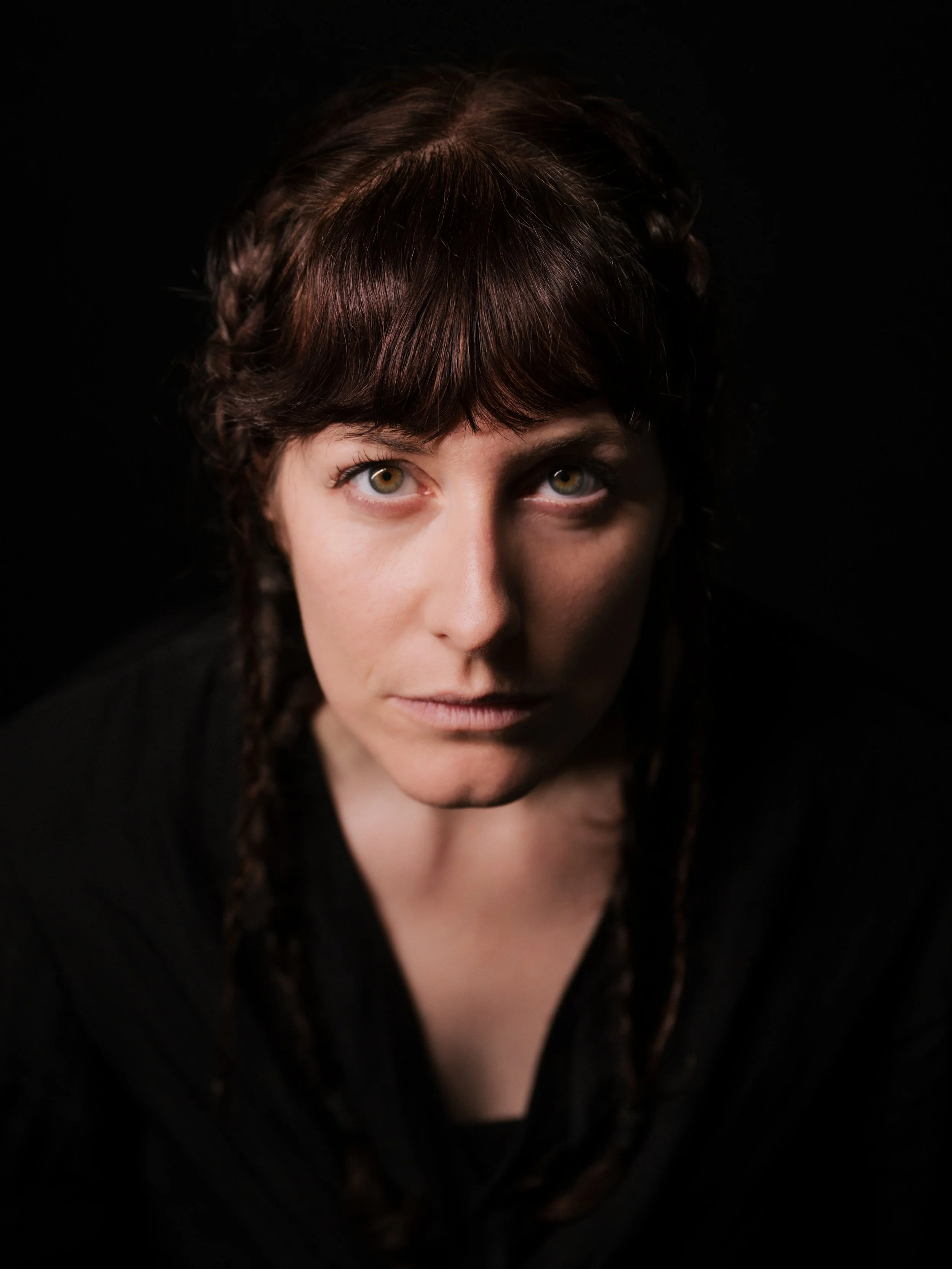 A woman with brown hair styled in braids, looking directly at the camera against a dark background.