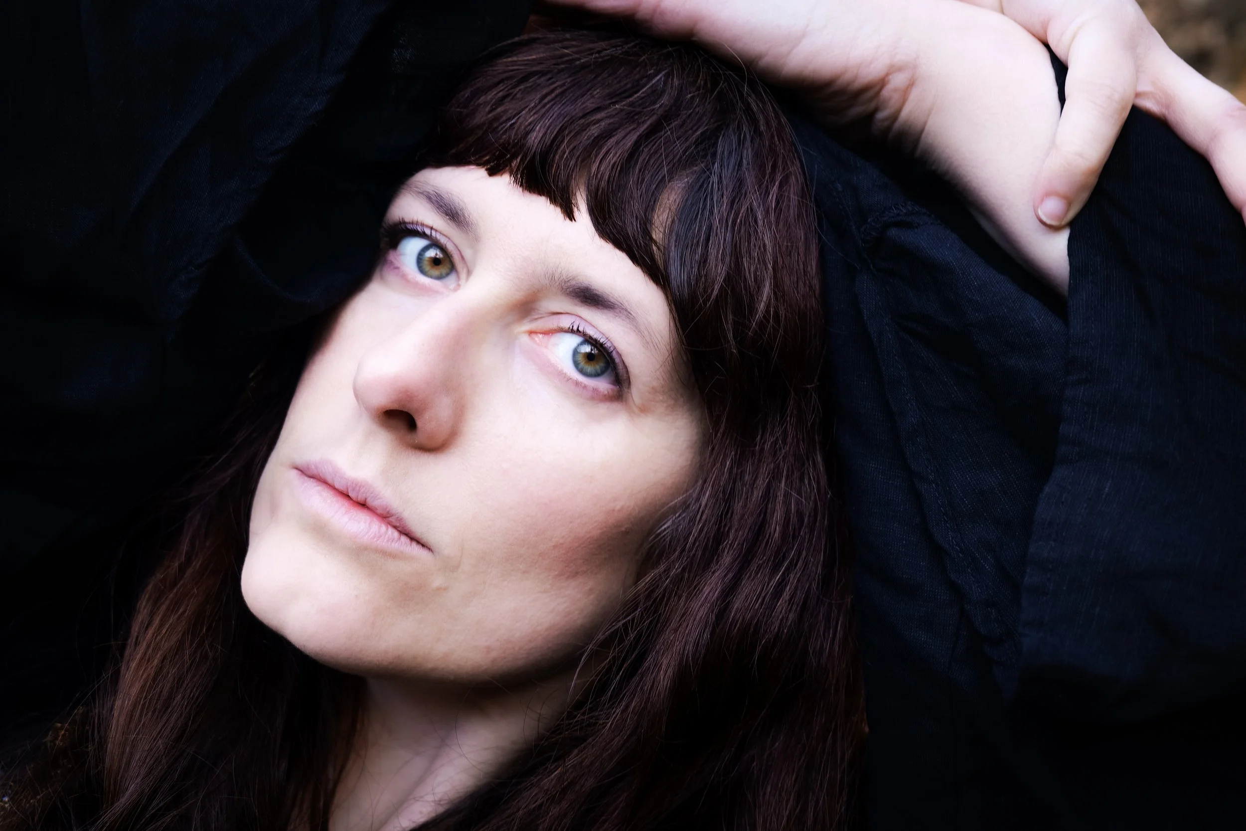 Close-up of a woman's face with long brown hair and hazel eyes, lying down with her head resting on her hand, wearing a black top.