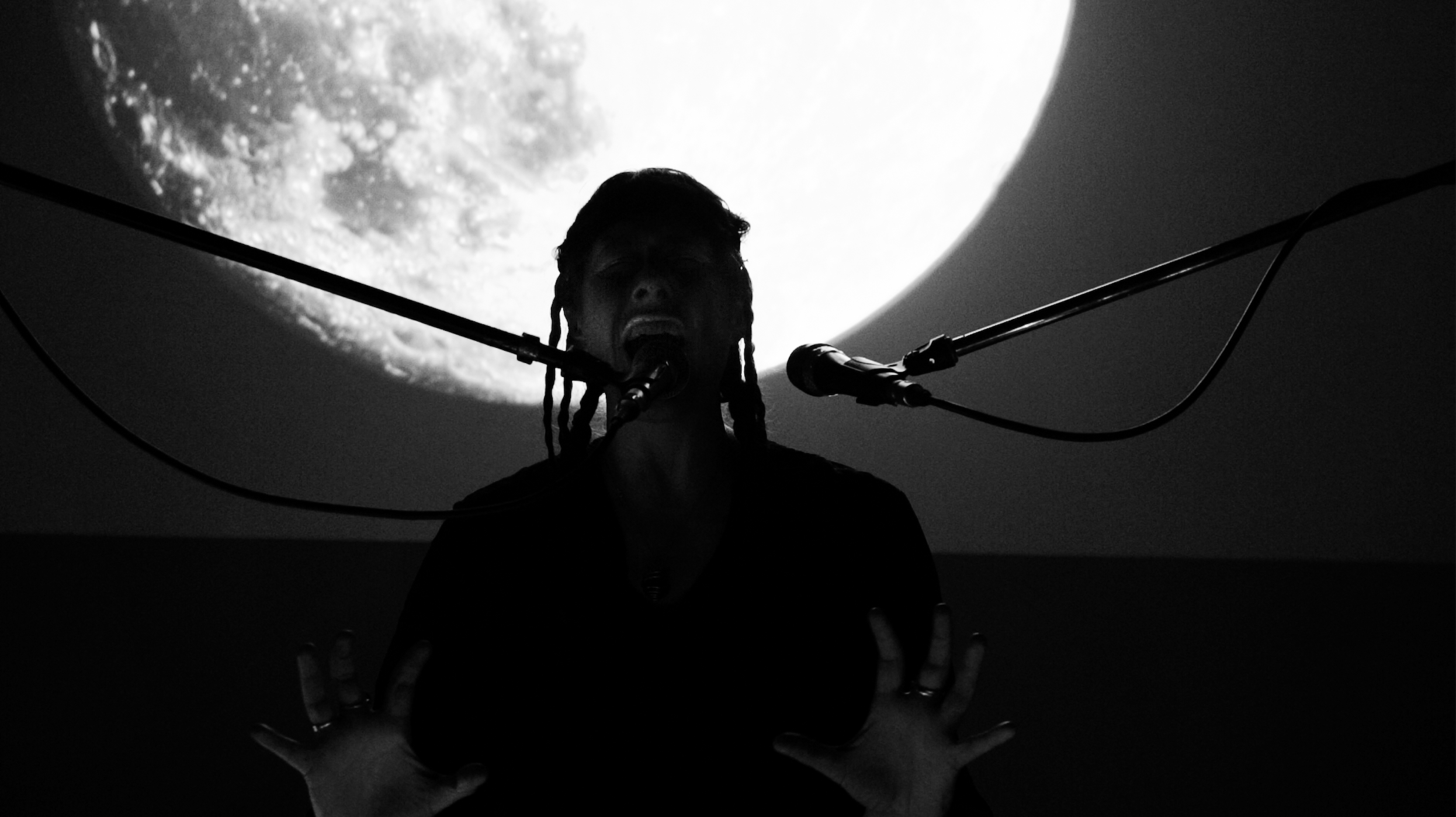 A person with dreadlocks singing into a microphone with their hands raised, silhouetted against a large illuminated moon in the background.